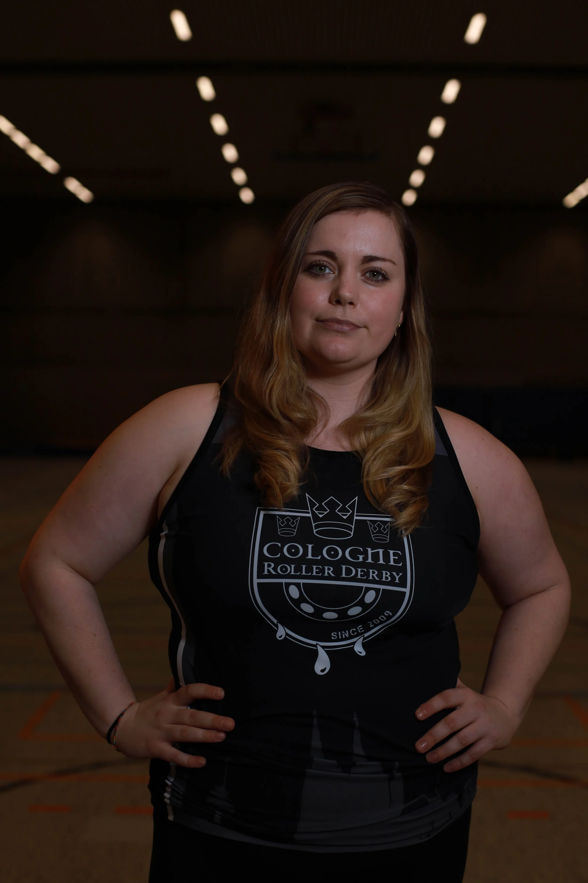 Eine junge Frau trägt ein schwarzes Trikot mit dem Logo des Kölner Roller Derby Teams und steht in einer Turnhalle.