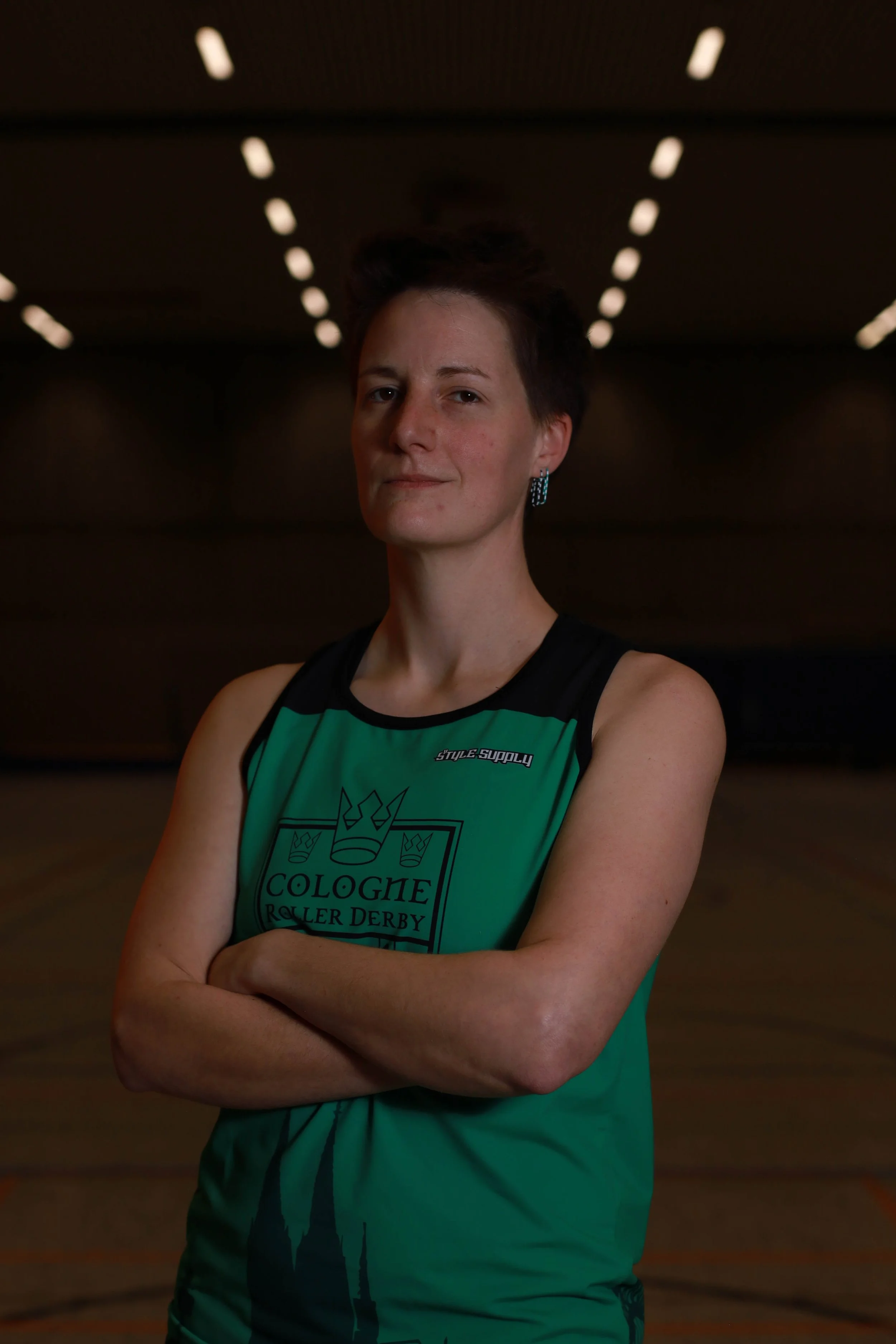 Ein selbstbewusst wirkendes weibliches Rollerderby-Teammitglied in einem grünen Trikot mit schwarzen Akzenten, das die Arme verschränkt in einer Halle mit dunklem Hintergrund steht.