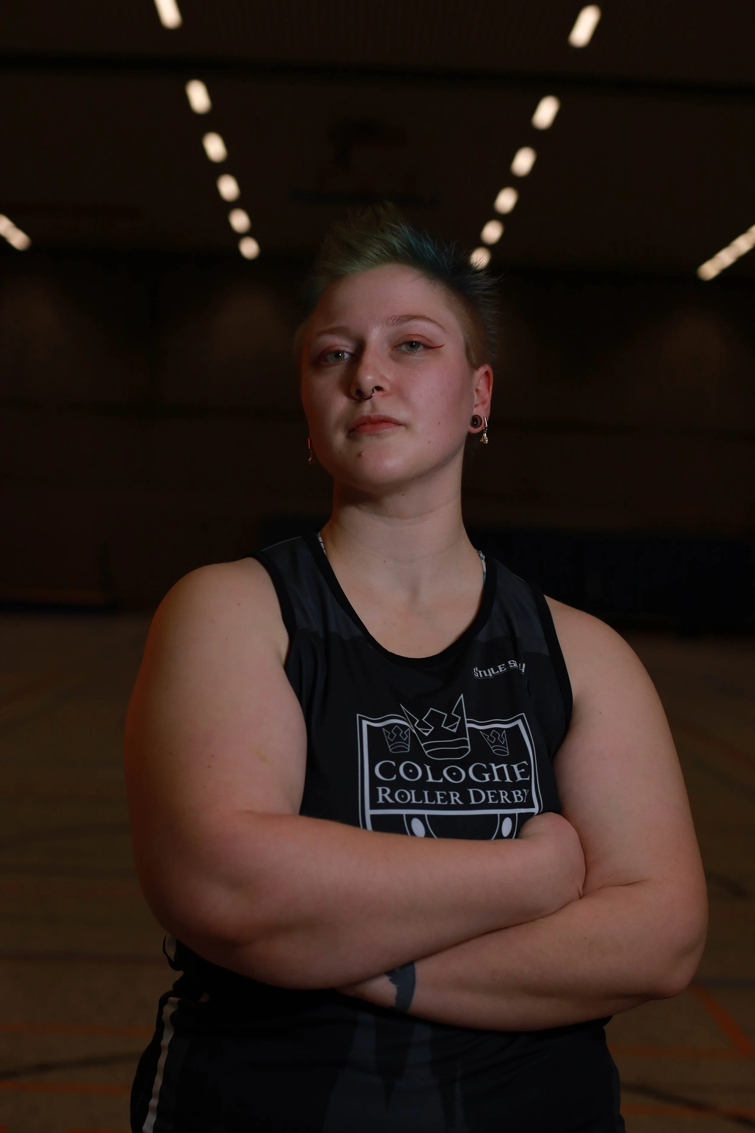Junge Person mit kurzen, gestylten Haaren und Piercings, steht in einer Turnhalle mit verschlüsseltem Blick, trägt ein schwarzes Sporttop mit einem Logo.