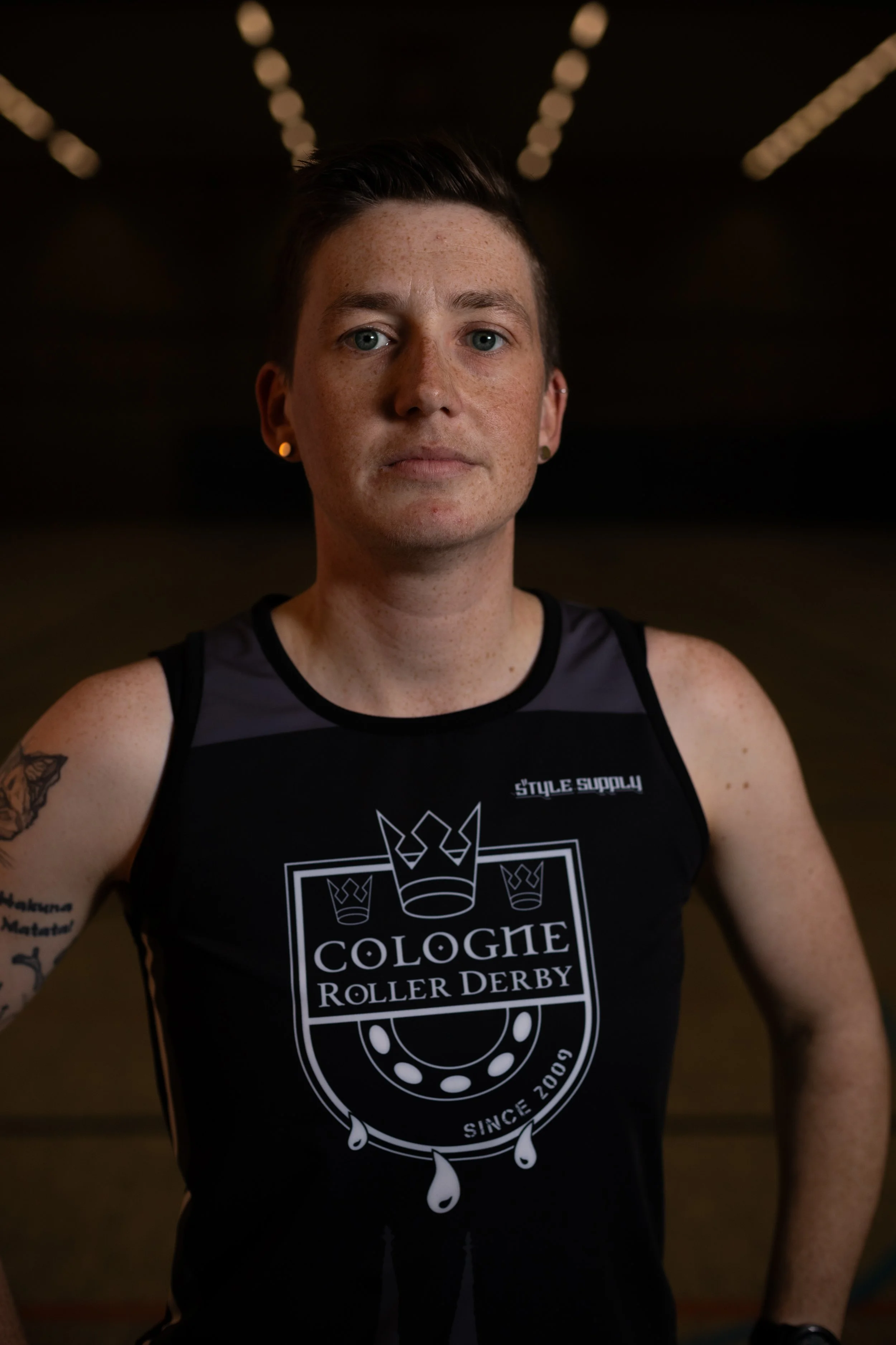 Junger Mann in einem schwarzen Trikot mit dem Logo "Cologne Roller Derby", im Hintergrund eine dunkle Halle mit künstlichen Lichtern