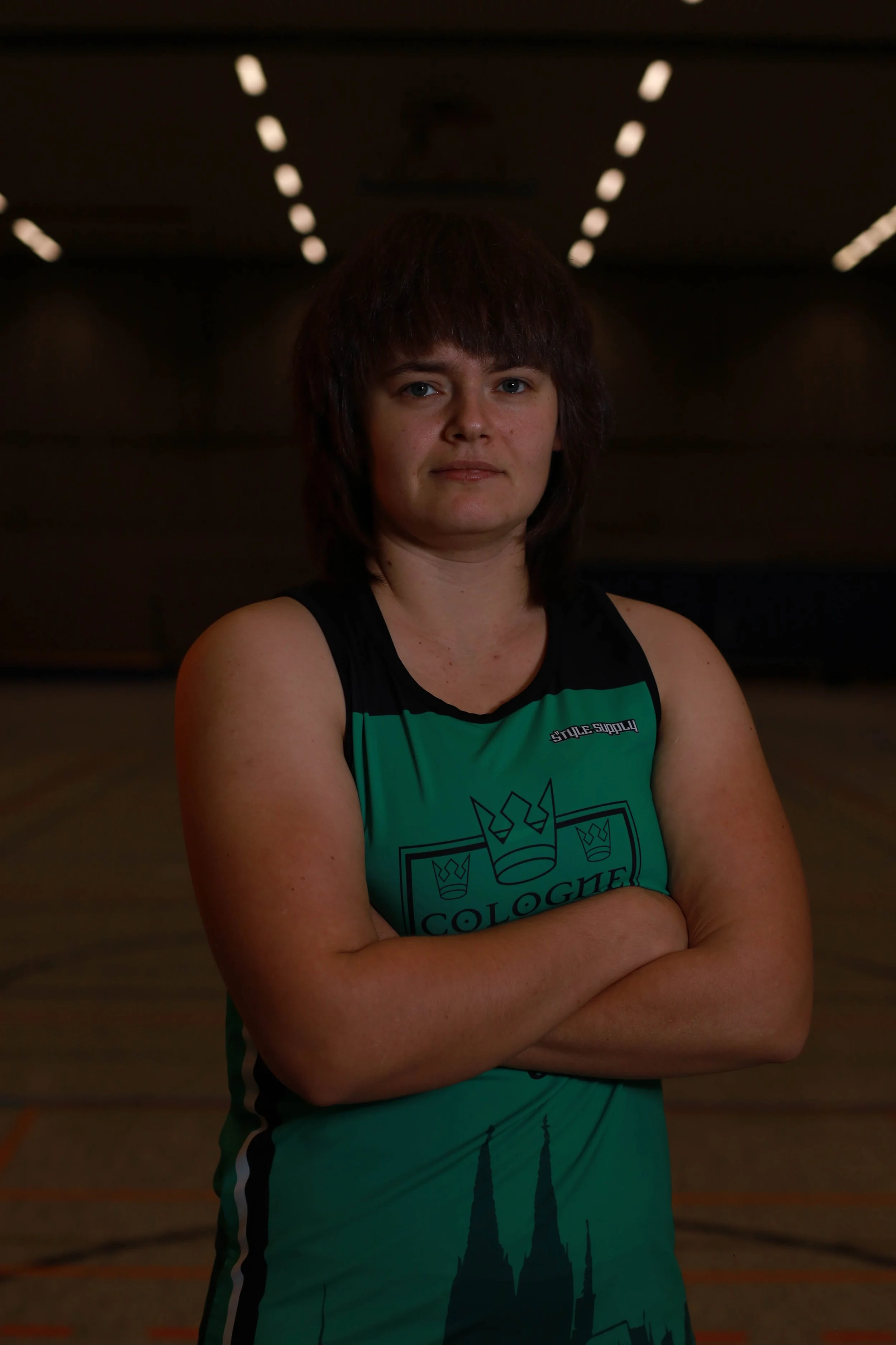 Junge Frau in einem grünen Sportdress steht in einem dunklen Sporthallenraum mit verschränkten Armen.