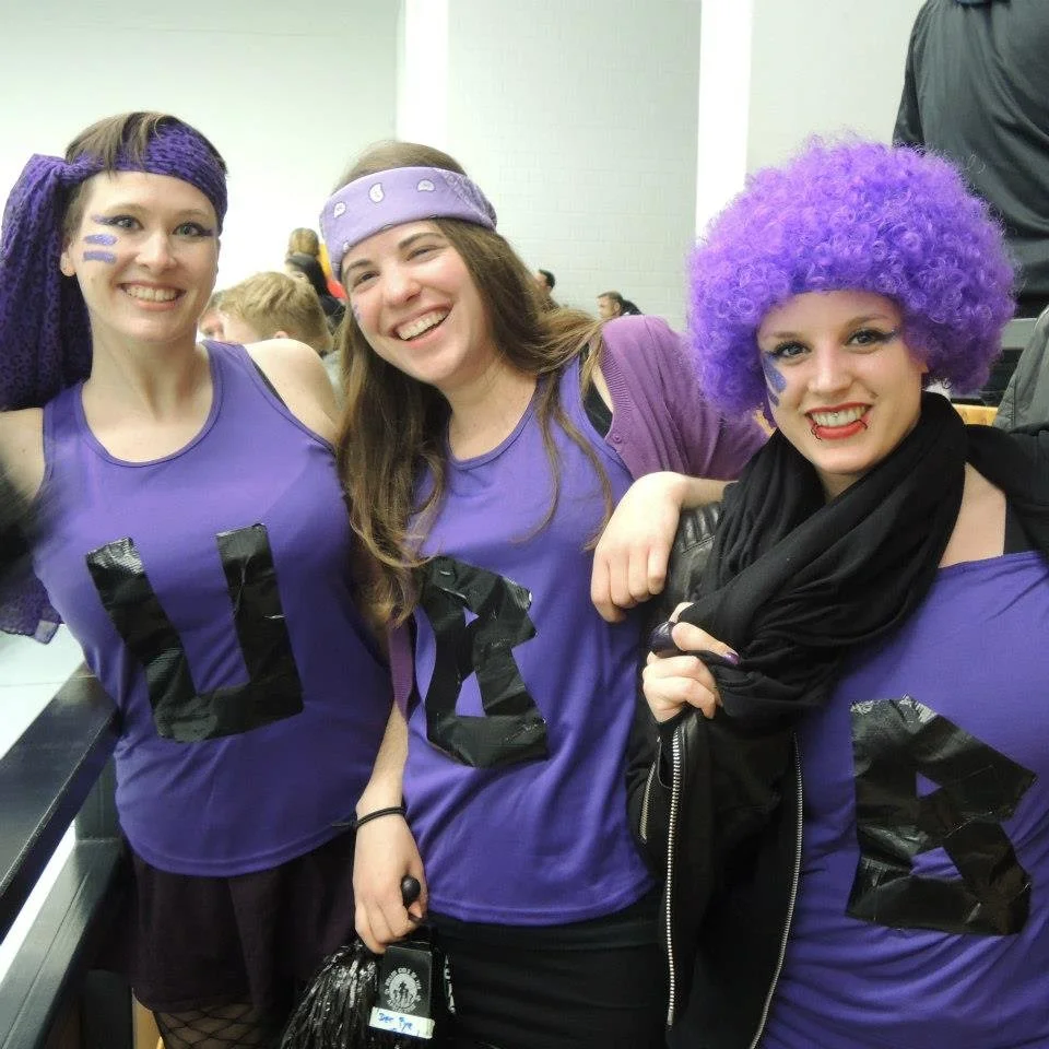 Drei Frauen in lila Trikots und Kostümen, die bei einer Veranstaltung lachen. Zwei tragen lila Haare, eine hat violette Haarsträhnen und trägt einen lila Lockenkopf und eine andere ein lila Stirnband. Sie scheinen Spaß zu haben.