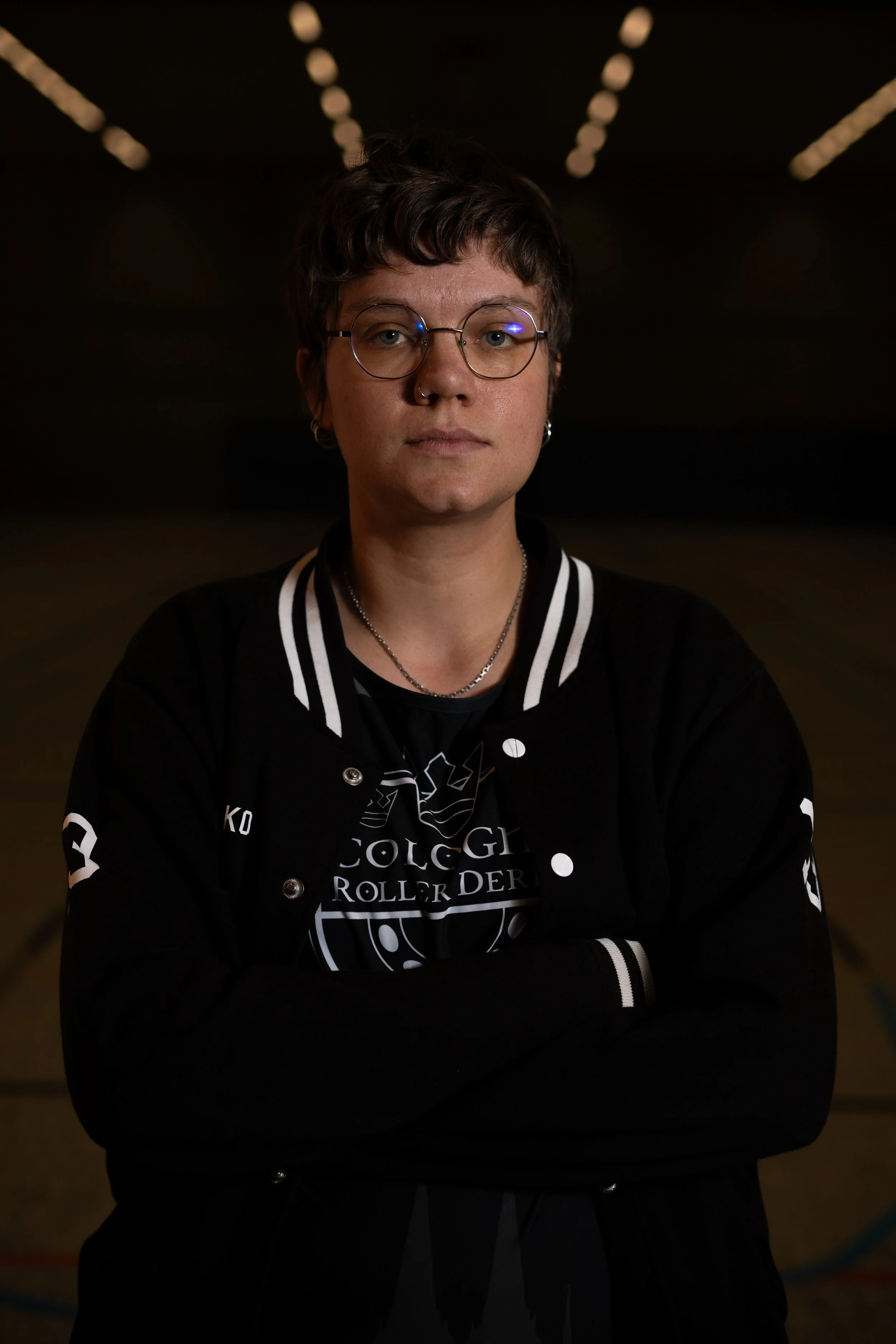 Porträt einer jungen Frau mit kurzen dunklen Haaren, runder Brille, schwarzen Jacke mit weißen Streifen, Armen verschränkt, im Dunkeln mit Lichtern im Hintergrund.