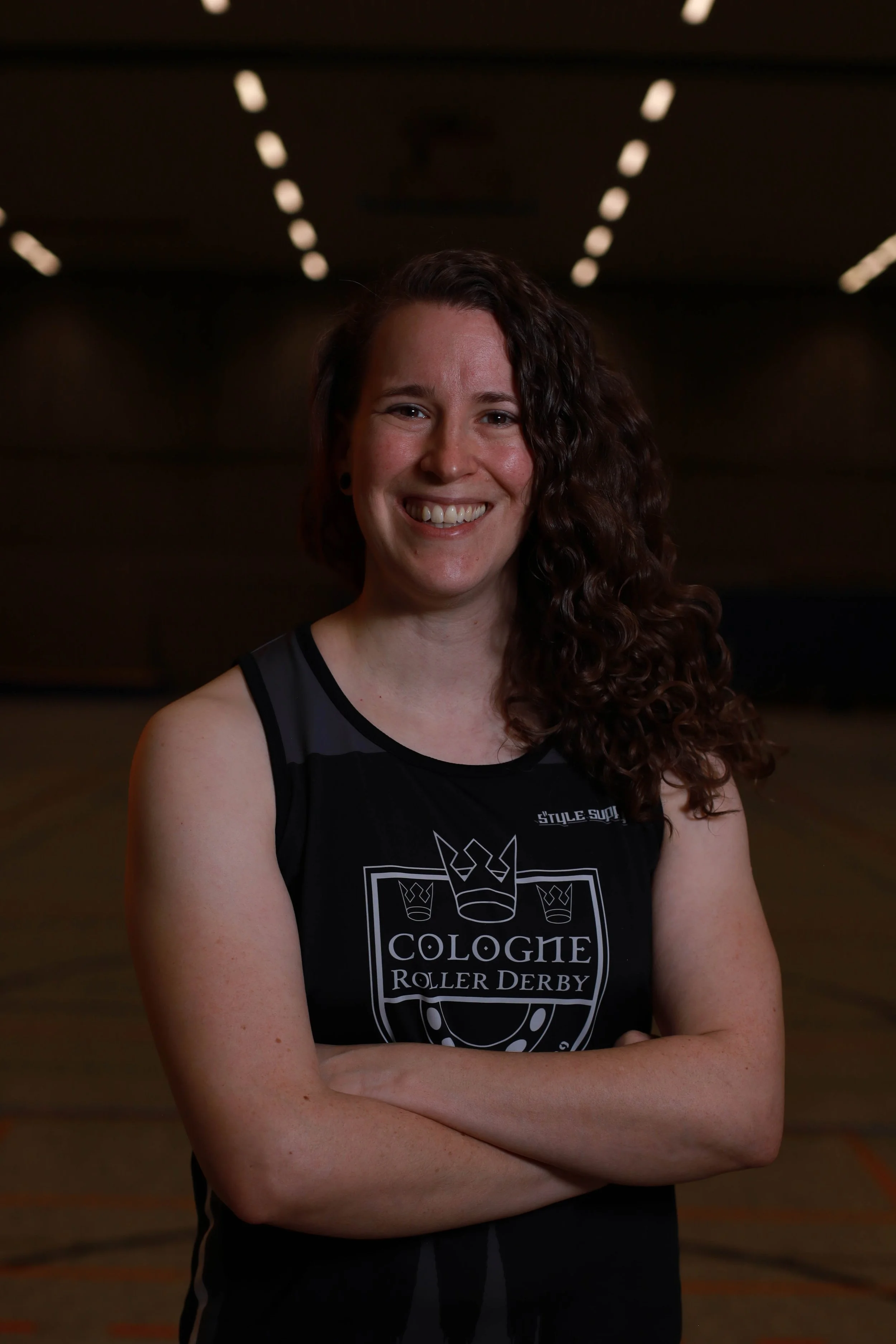 Eine lachende Frau mit lockigem braunem Haar in einem schwarzen Trikot mit dem Aufdruck "Cologne Roller Derby" steht in einer Rollschuhhalle.