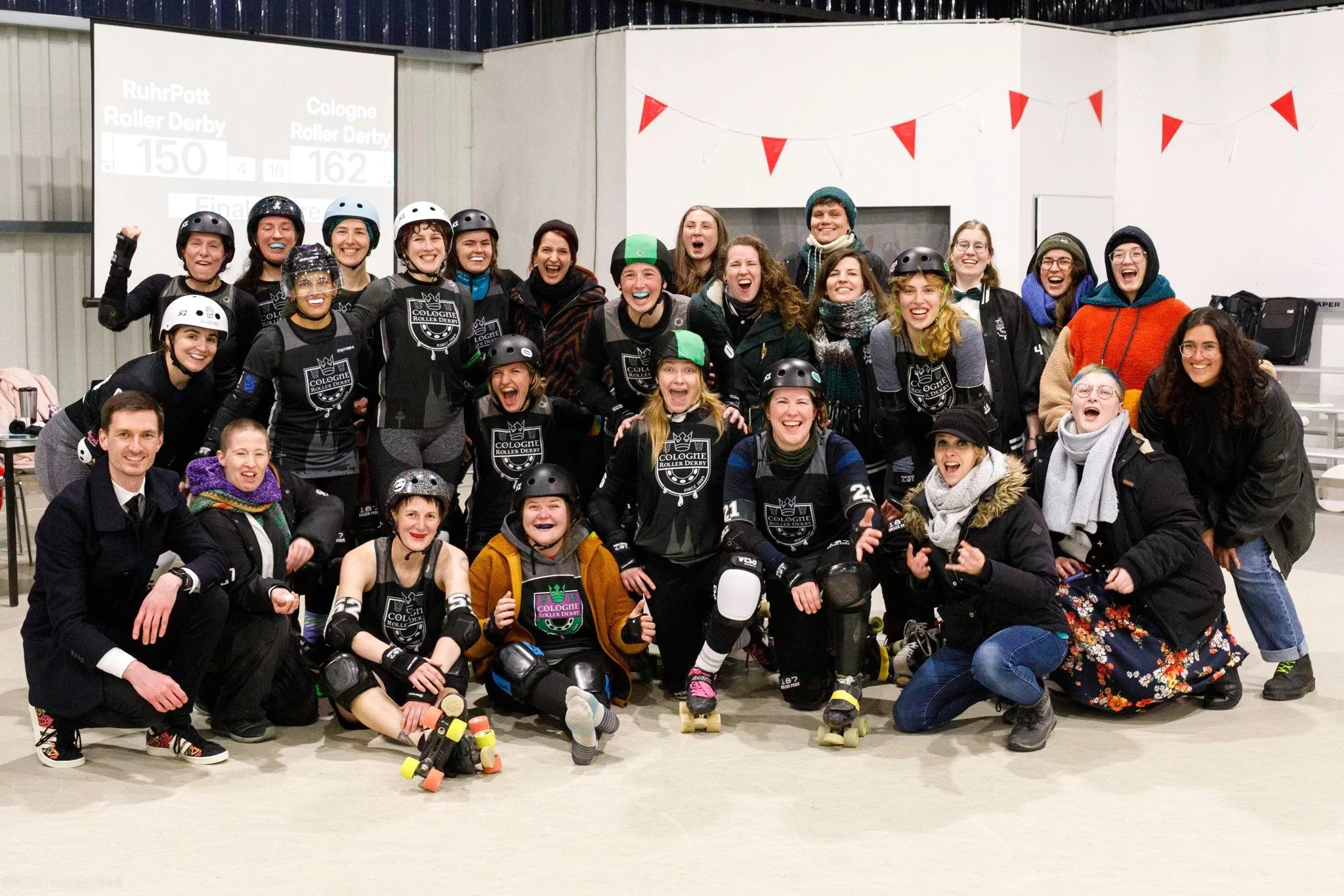 Eine Gruppe von Menschen, darunter Rollschuhfahrerinnen in Schwarz mit Helmen und Schutzausrüstung, die nach einem Rollschuhwettbewerb in einer Halle glücklich zusammenstehen und jubeln.