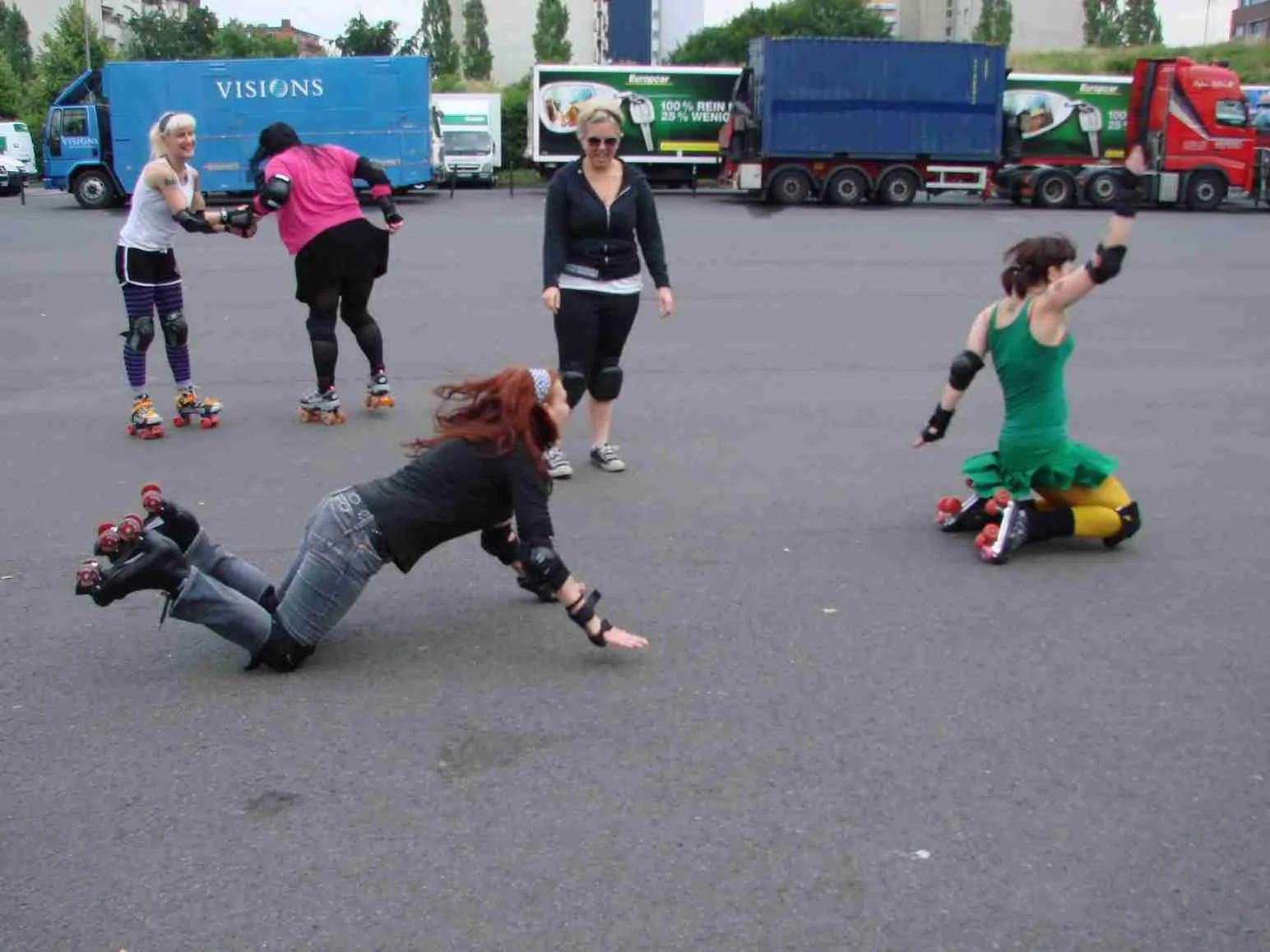 Vier Frauen beim Rollschuhlaufen auf einer asphaltierten Fläche, eine hilft einer anderen beim Aufstehen, im Hintergrund stehen drei weitere Frauen, zwei in Spielzeugmode, alle tragen Schutzausrüstung.