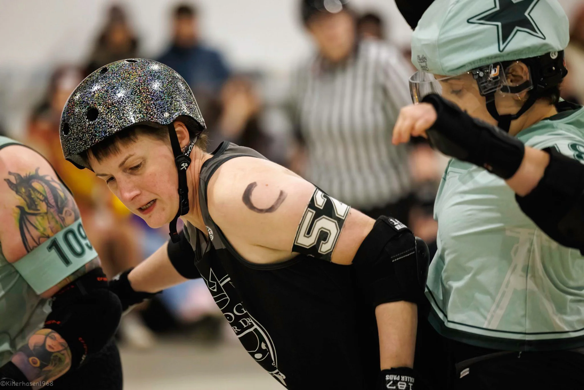 Roller Derby Spielerin mit einem durchscheinenden Helm und Tattoos auf dem Arm, die gegen eine andere Spielerin kämpft, während ein Schiedsrichter im Hintergrund beobachtet.