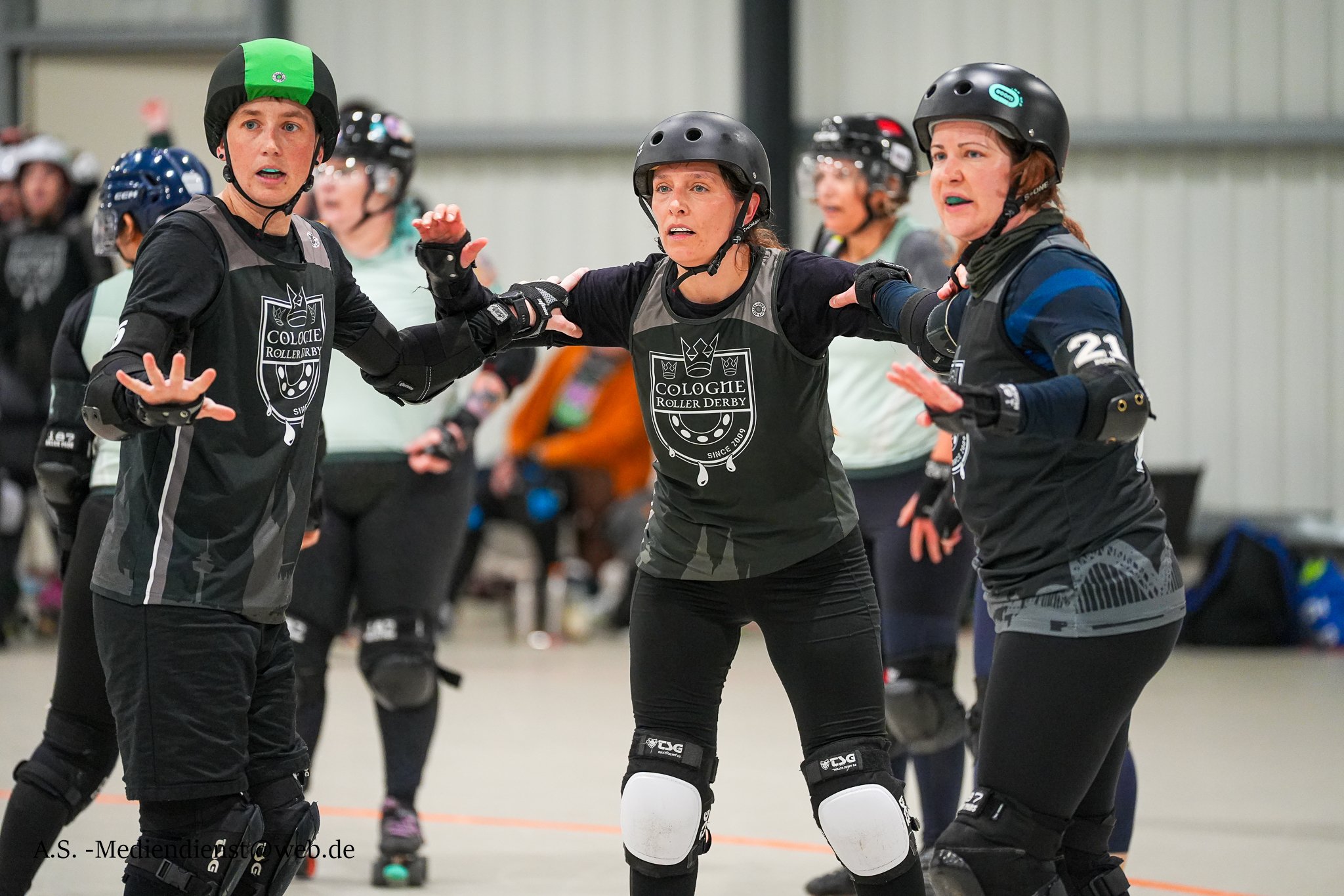 Drei Rollerderby-Spielerinnen in Schutzausrüstung während eines Spiels in einer Halle, einige andere Spieler im Hintergrund.