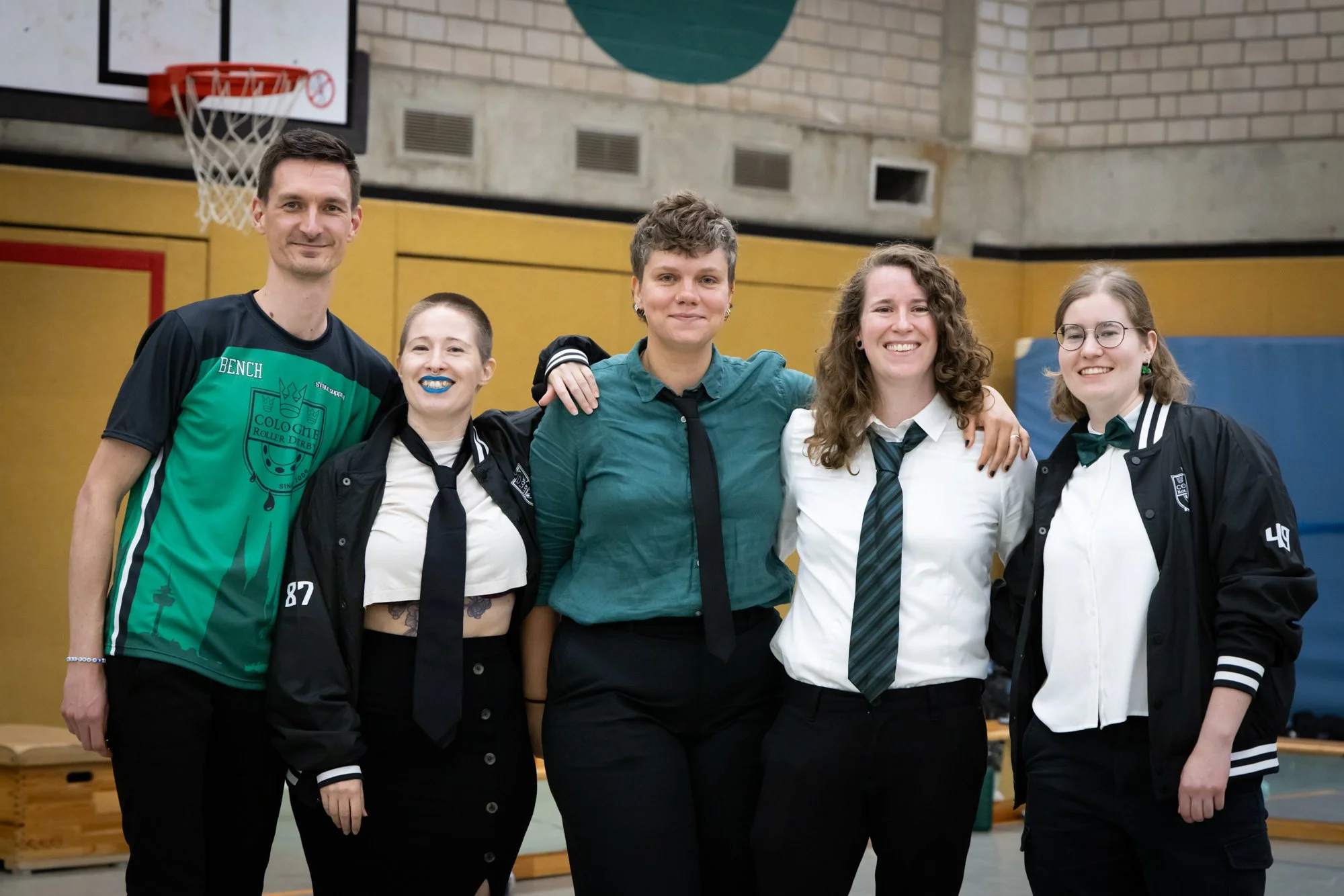 Gruppenfoto von fünf jungen Erwachsenen, die in einem Turnsaal stehen, mit lachenden Gesichtern und Schuluniformen, einige mit Krawatten, hinter einem Basketballkorb.