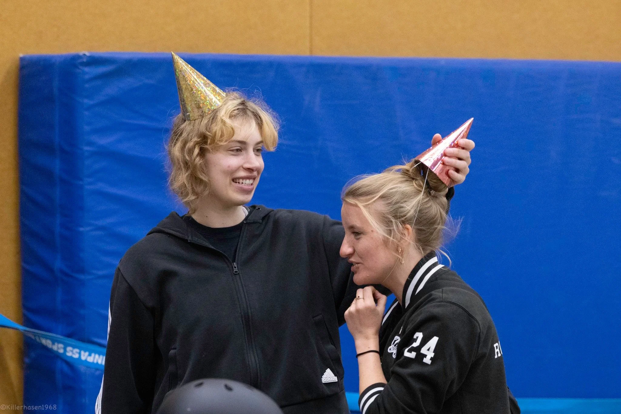 Zwei Frauen feiern mit Partyhüten, eine lächelnde Frau legt einem anderen jungen Frau Hut auf den Kopf, im Hintergrund blaue Matte