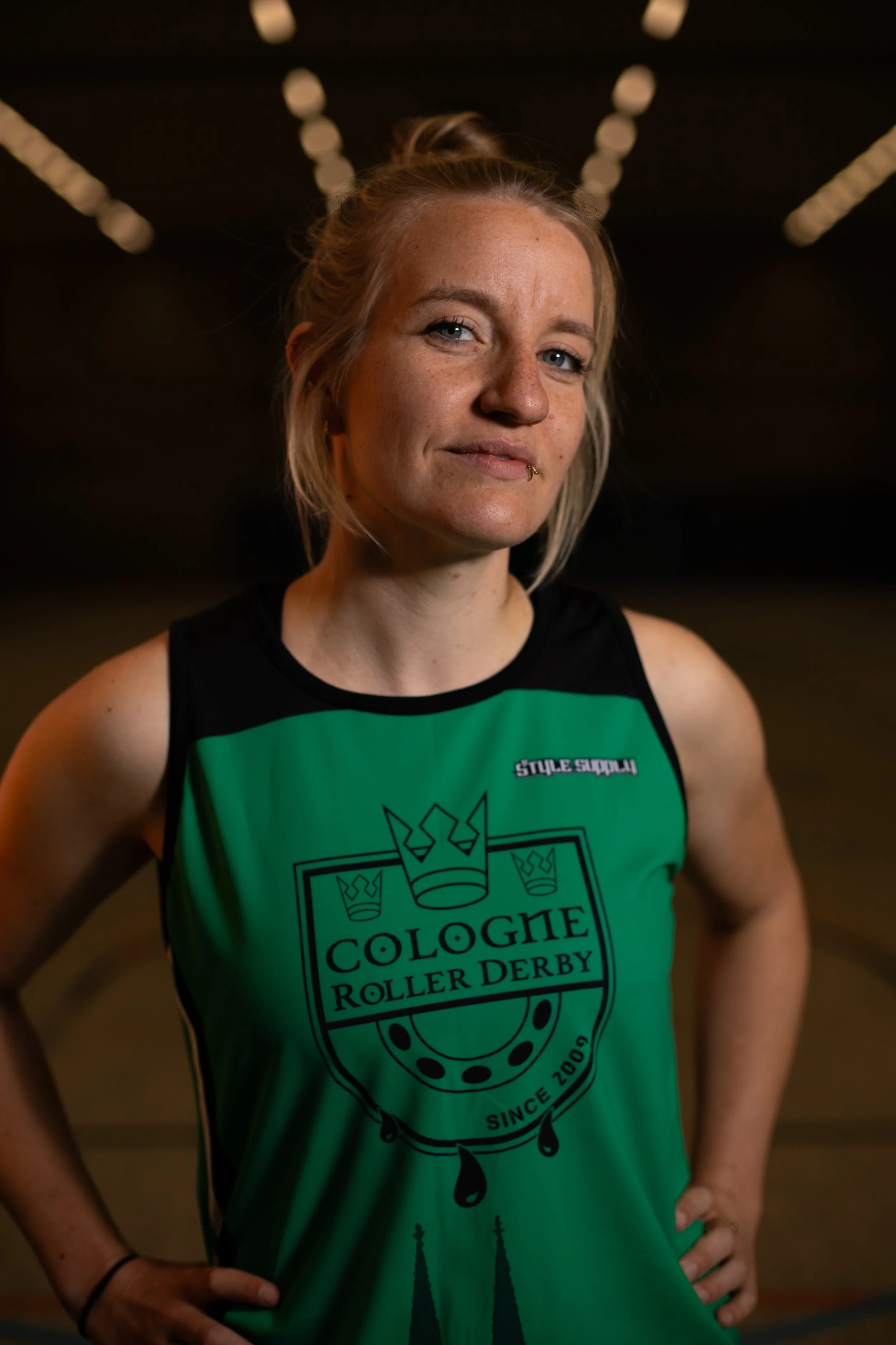 Eine Frau trägt ein grünes Trikot mit dem Logo 'Cologne Roller Derby' und steht in einer Turnhalle.