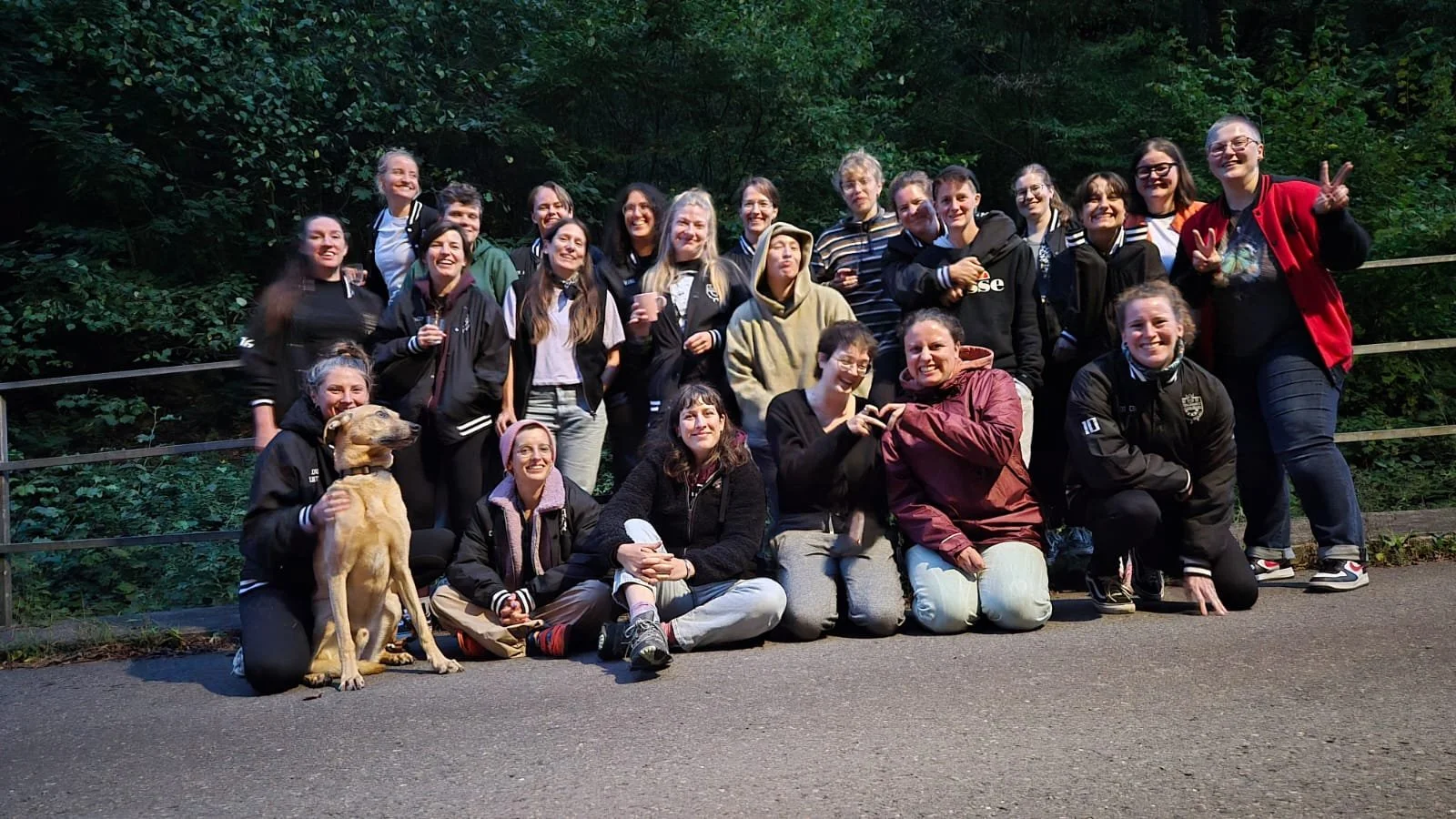 Gruppe von Frauen und einem Hund im Freien bei Sonnenuntergang, lächelnd und winkend.