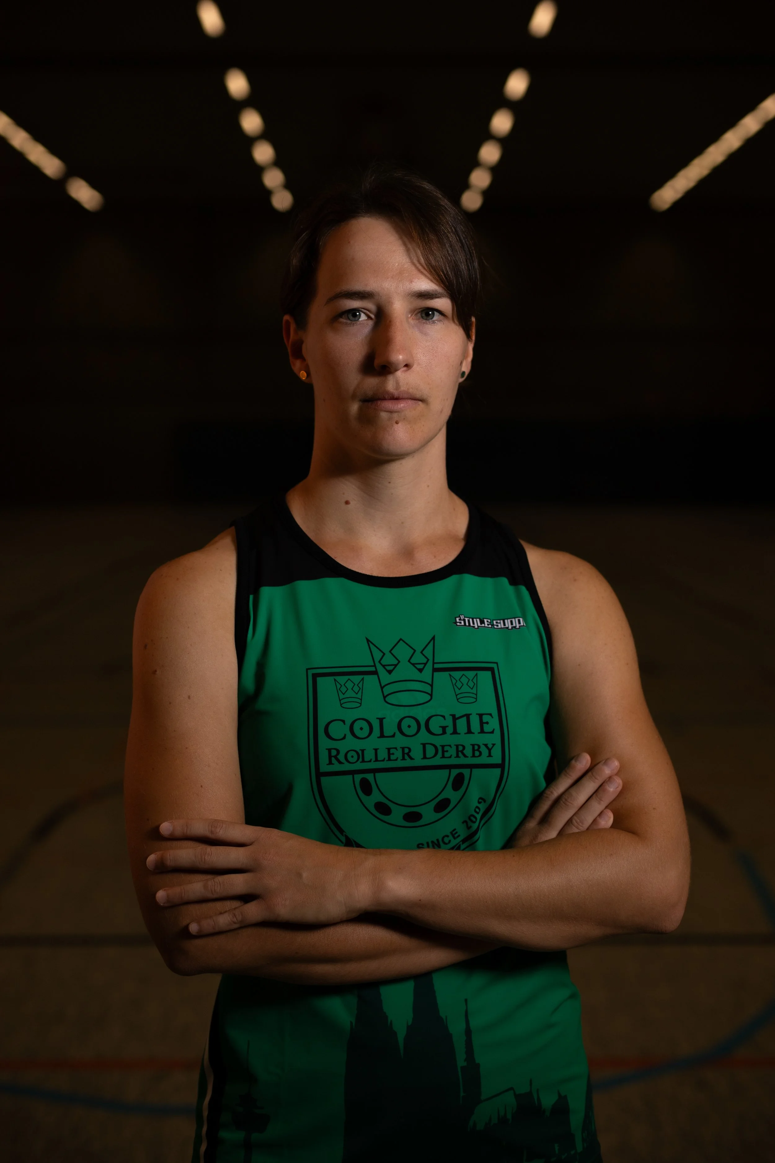 Eine Frau in einem schwarzen und grünen Rollschuh-Outfit steht in einer Turnhalle mit verschränkten Armen und Blick direkt in die Kamera.
