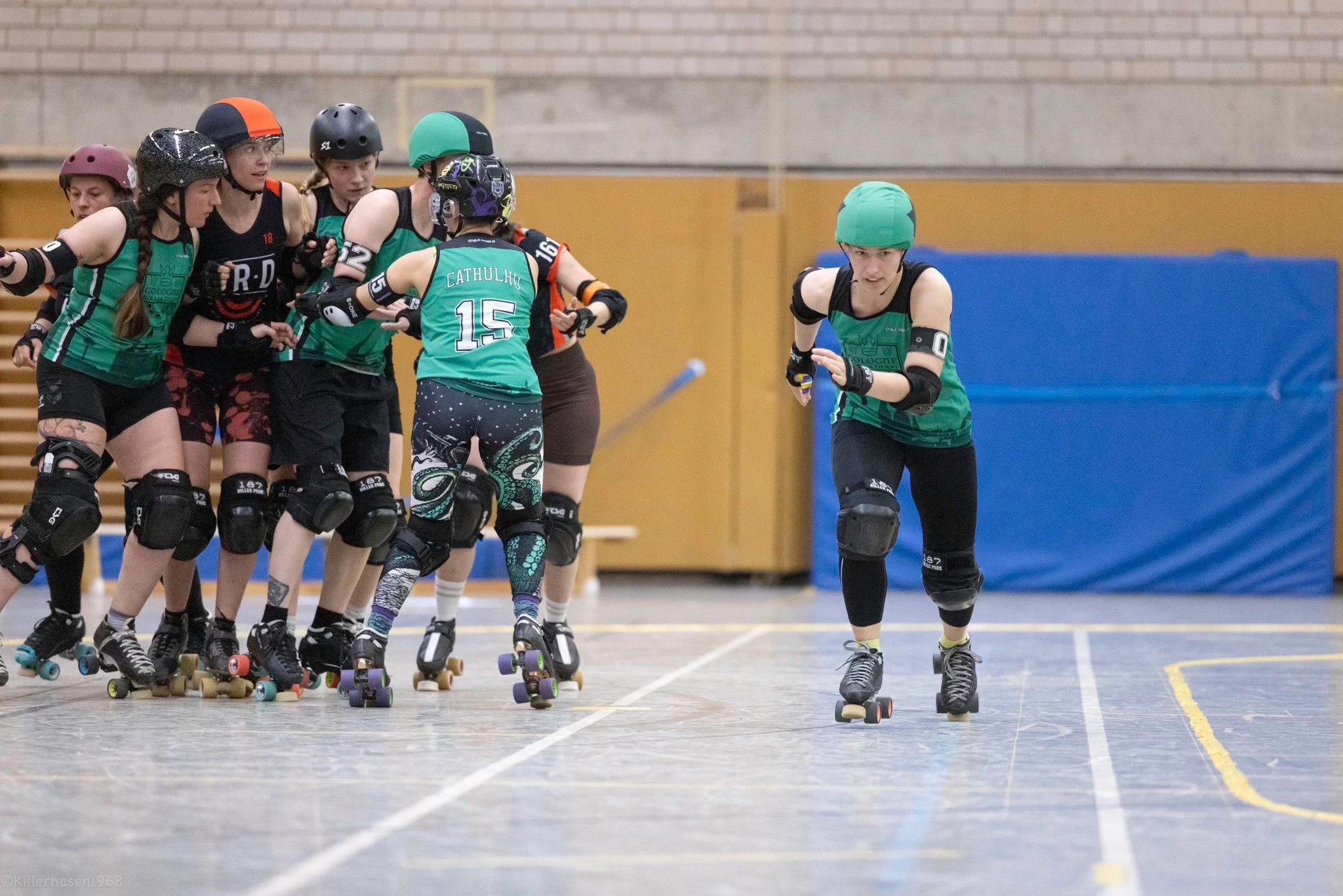 Rollschulfahrerinnen bei einem Wettkampf auf einer Rollschuhbahn, mit einer Person in grünem Trikot, die sich von der Gruppe löst.