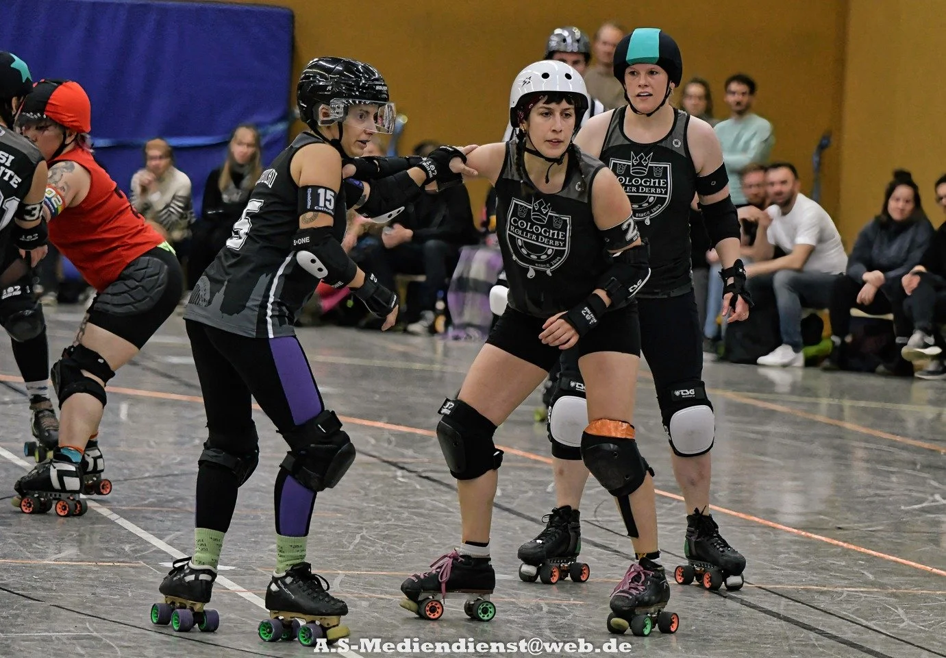 Drei Frauen beim Rollerderby, die im Team mit schwarzen Trikots und Helm spielen, eine weibliche Spielerin hilft einer anderen, beim Vier gegen Vier spielen. Im Hintergrund sitzen Zuschauer im Indoor-Sporthalle.