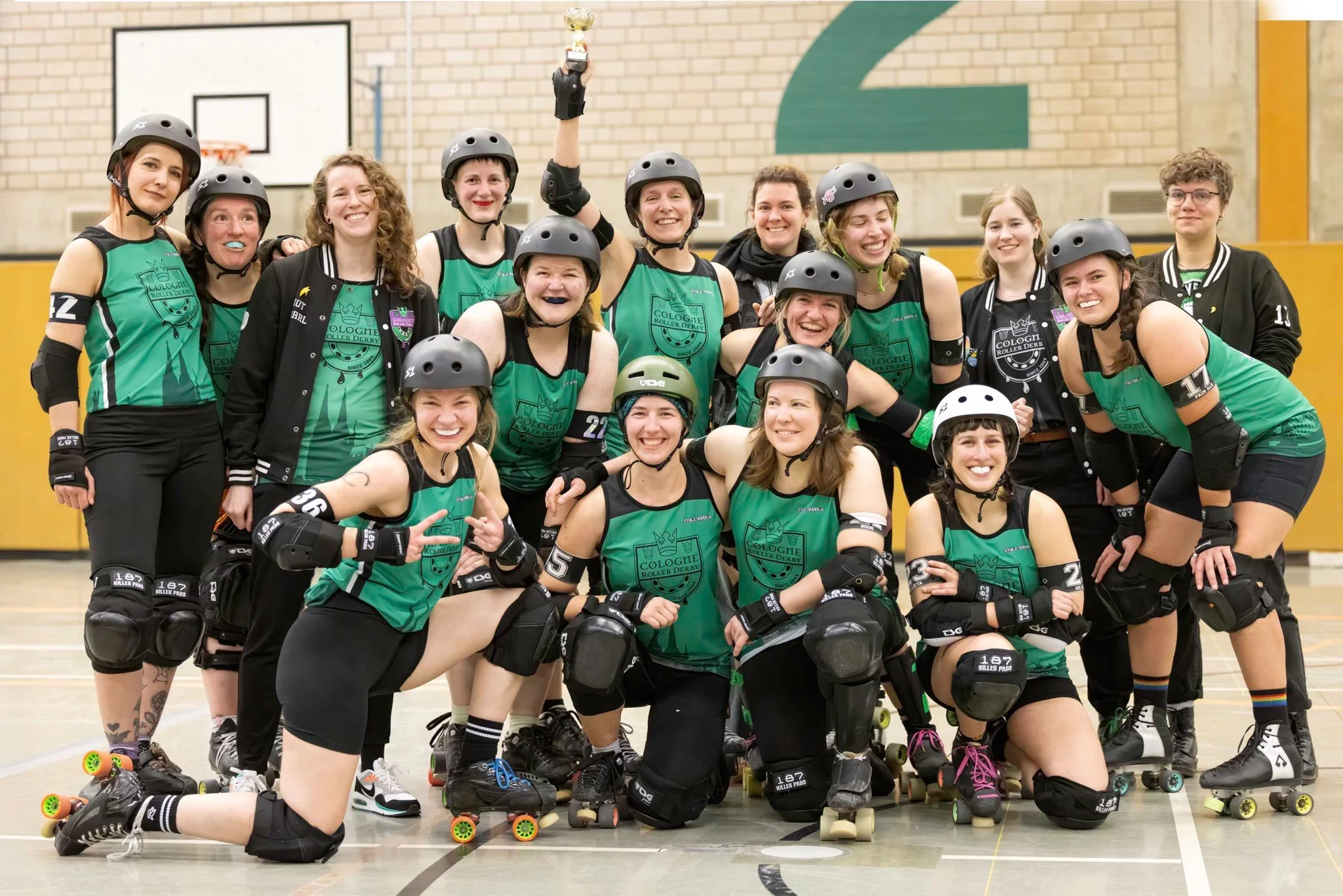 Gruppe von Frauen auf Rollschuhen, die nach einem Rollerderby-Spiel eine Siegestrophäe hochhalten, im Innenraum einer Sporthalle, alle tragen Schutzhelme und Sportkleidung in Grün und Schwarz.