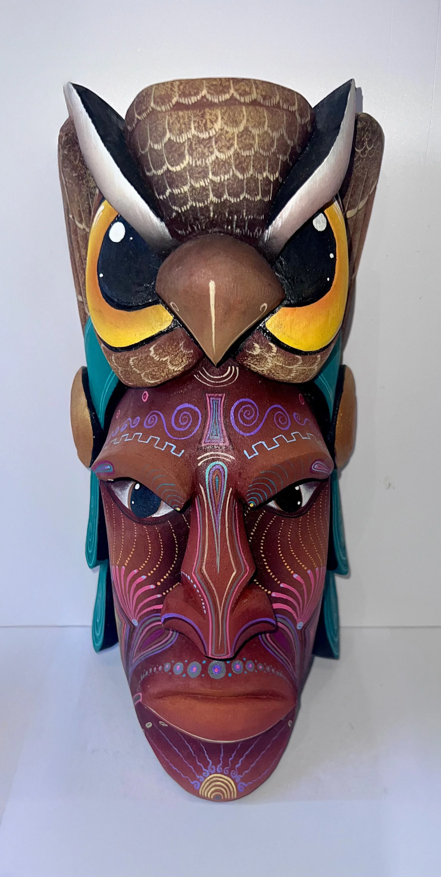 Colorful carved wooden mask with intricate patterns, depicting a mystical face with large eyes, a prominent nose, and a headdress featuring an owl.