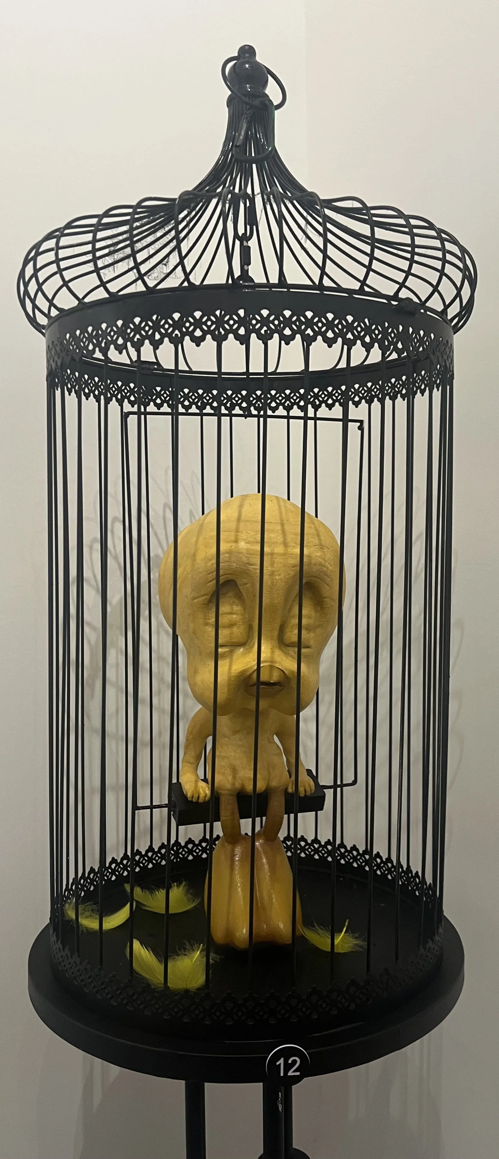 Yellow wooden sculpture of a large-headed, small body humanoid figure inside a black decorative birdcage with yellow feathers on the floor.