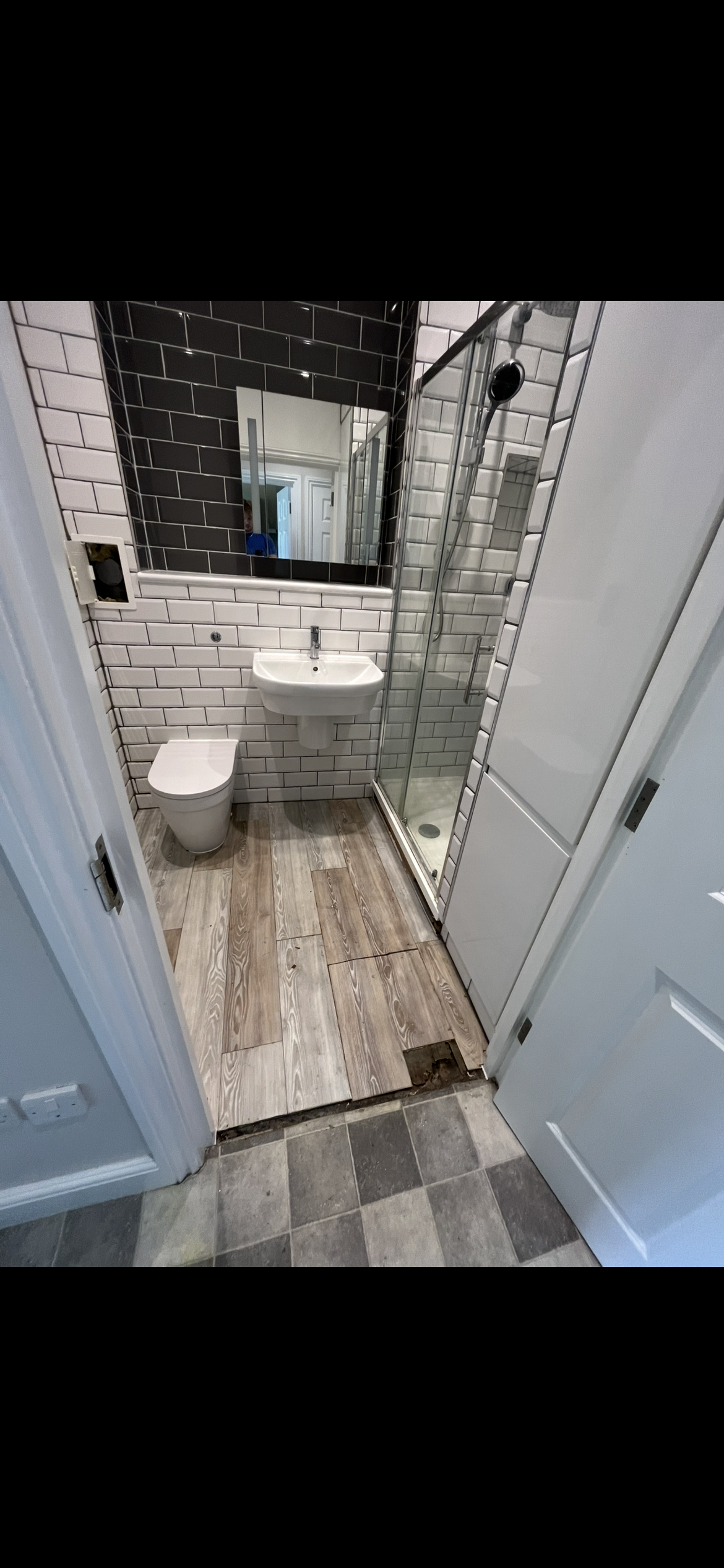 Small bathroom with white subway tile walls, black accent tiles, a wall-mounted sink, a toilet, a shower with a sliding glass door, and wood-like flooring partially removed revealing a subfloor.