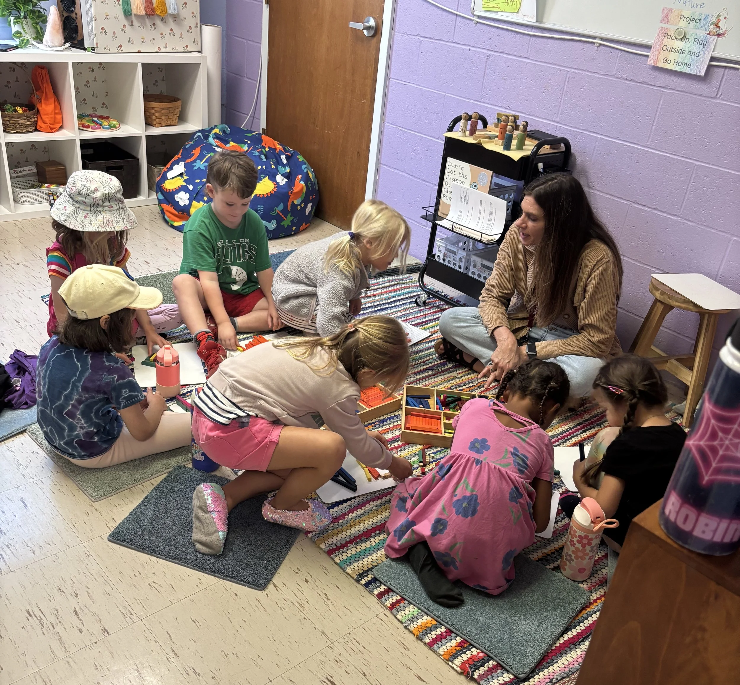 A teacher leaning over a table to assist a young girl with blonde hair as she writes on a piece of paper. The classroom has purple walls, chairs, and educational decor, including a colorful sign on the bulletin board that says, 'This is where the fun stuff happens.'