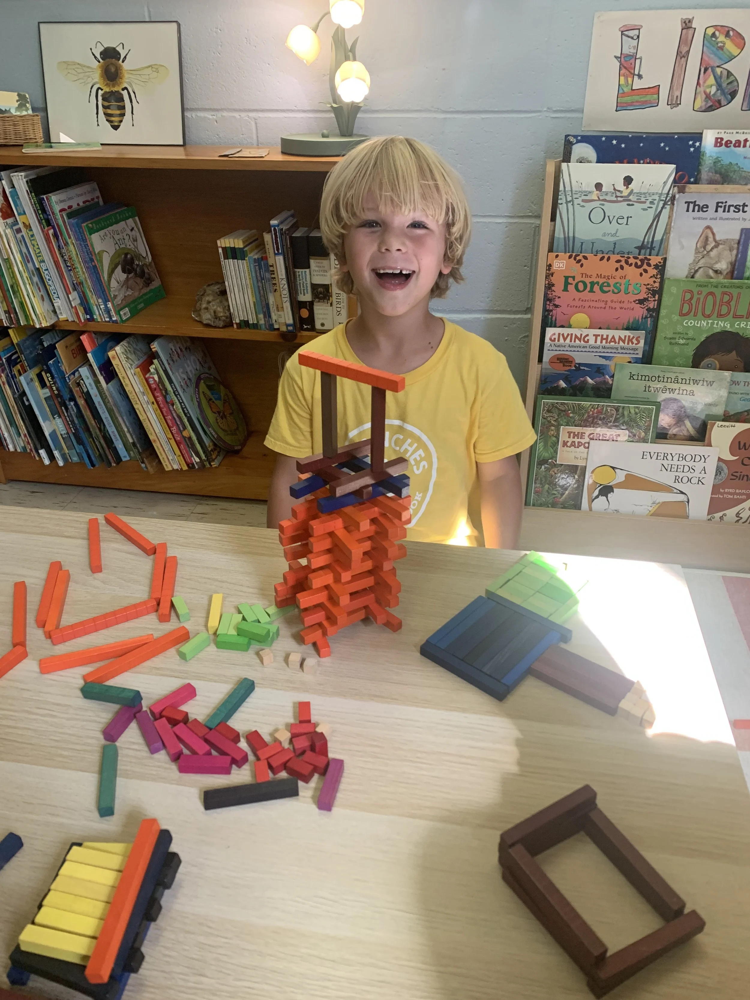 A young boy with blonde hair and a yellow shirt is smiling while sitting at a table with colorful wooden block structures and loose blocks, in a room with bookshelf and bookshelf with children’s books in the background.