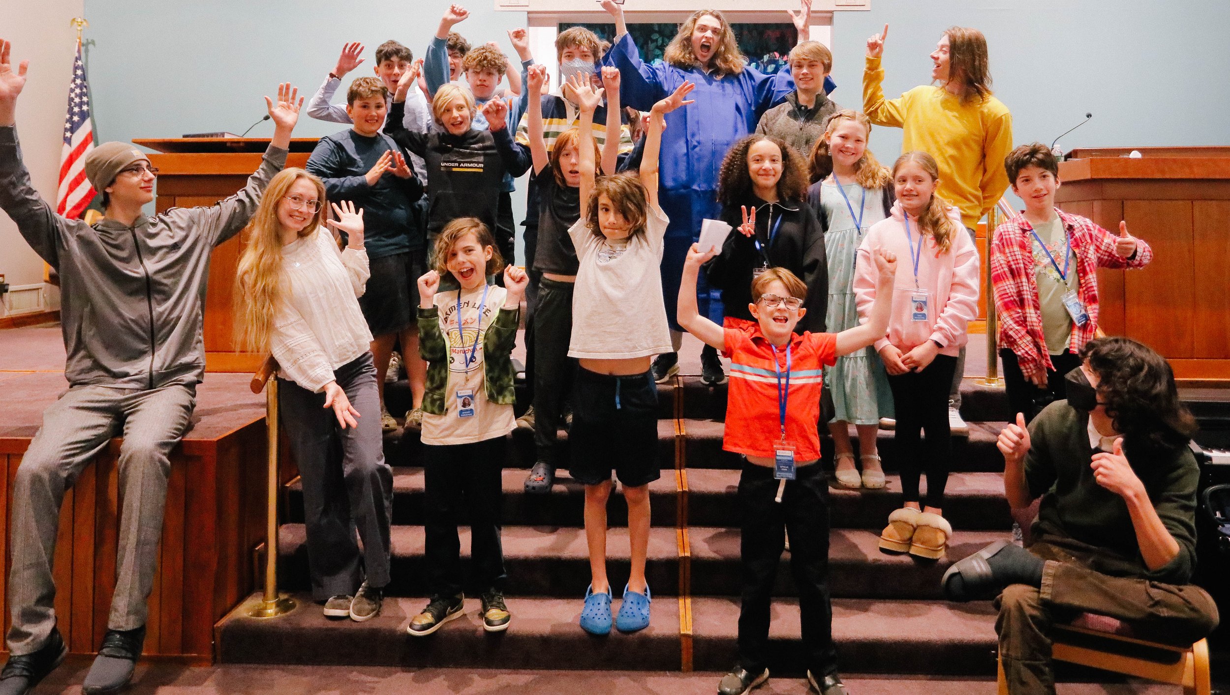 Group of children and two adults celebrating on a stage, smiling, raising their hands, and making peace signs, with an American flag in the background.