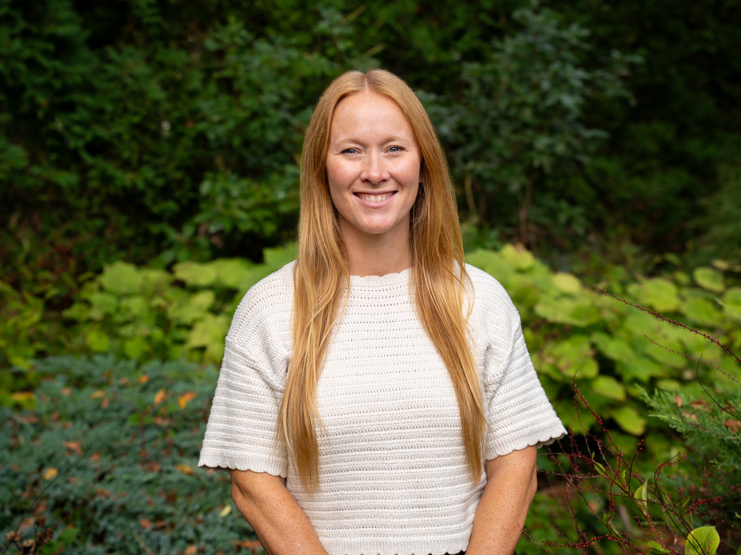A woman with long red hair and fair skin smiling outdoors, wearing a white knitted sweater, with green leafy plants in the background.