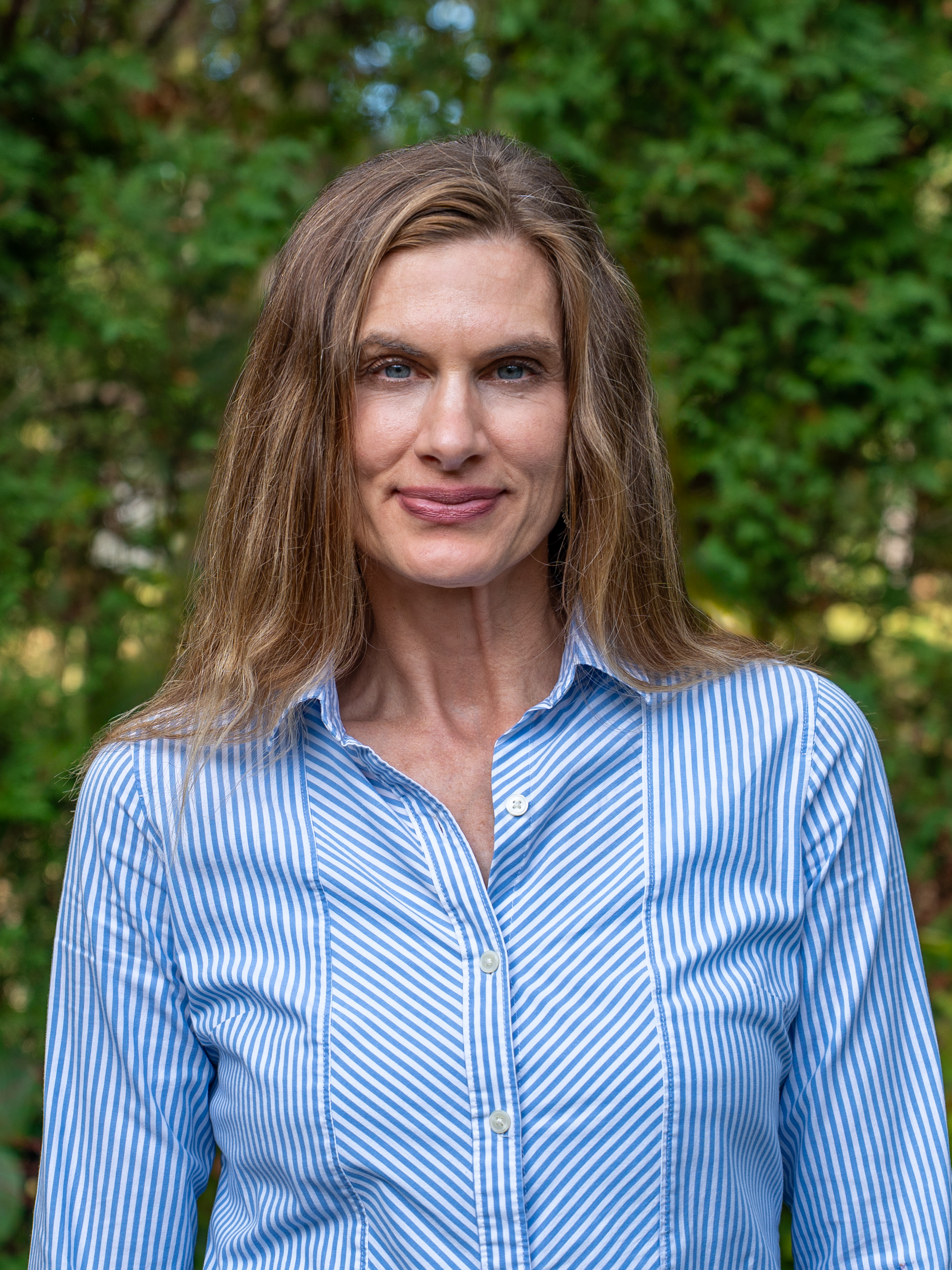 A woman with long light brown hair and blue eyes wearing a blue and white striped button-up shirt outdoors with green foliage in the background.