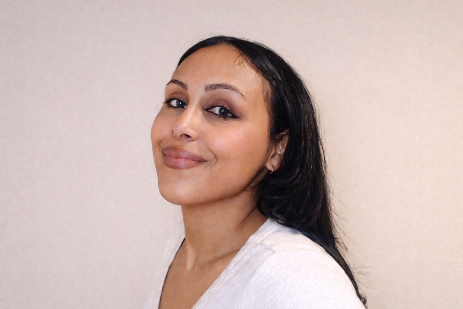 A woman with dark hair, light makeup, and a white shirt, smiling slightly and looking over her shoulder in front of a plain beige wall.