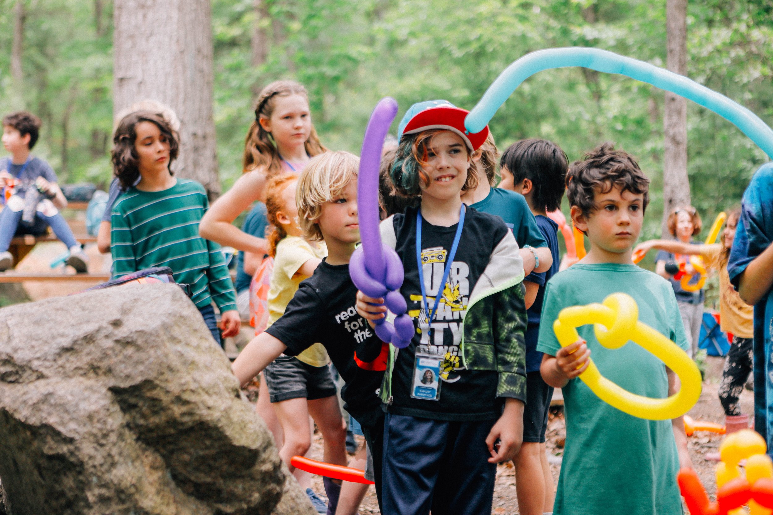 Group of children outdoors in a wooded area, holding colorful balloon sculptures during a daytime event.