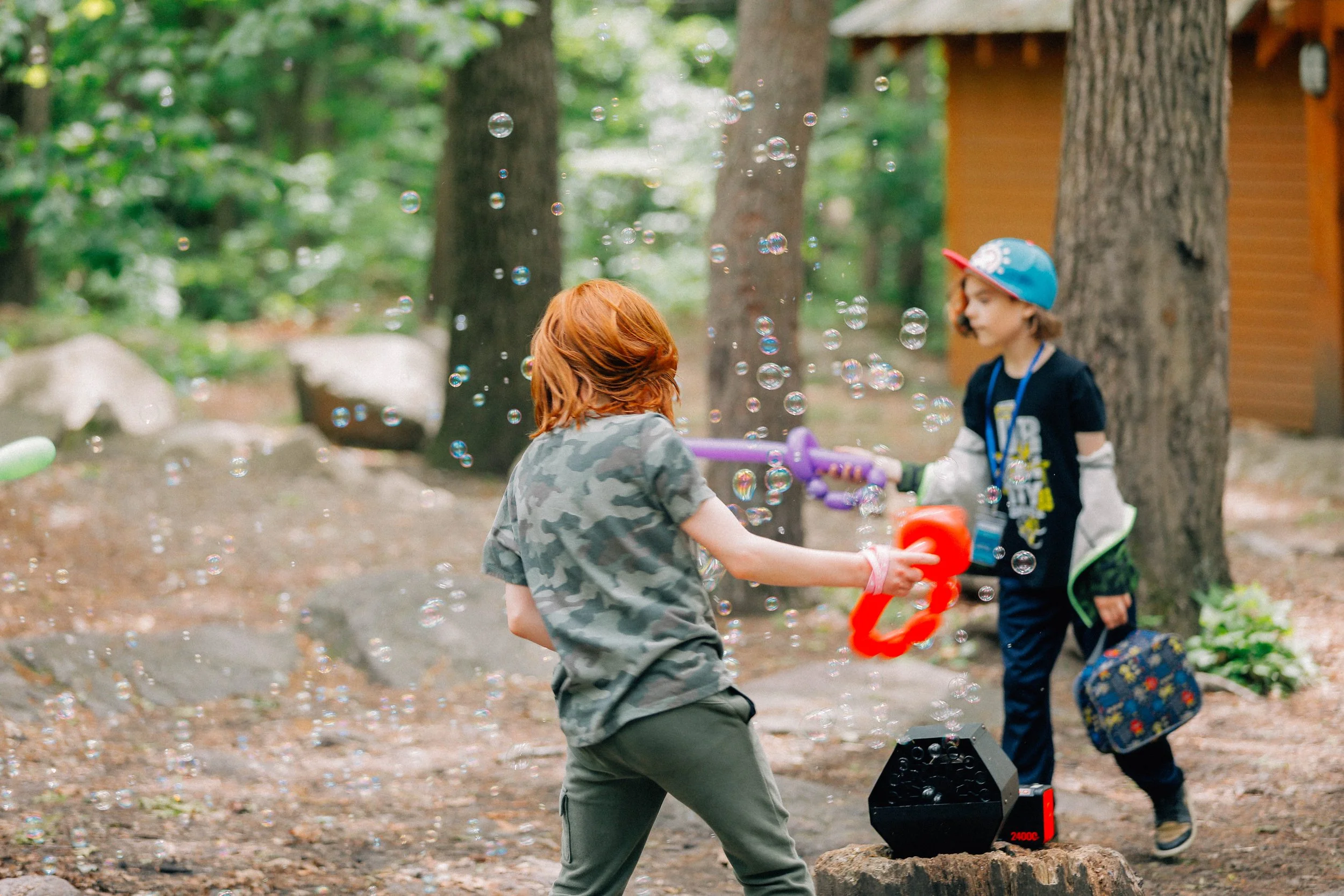 Two children playing with bubbles and water guns in a wooded outdoor area during daytime.