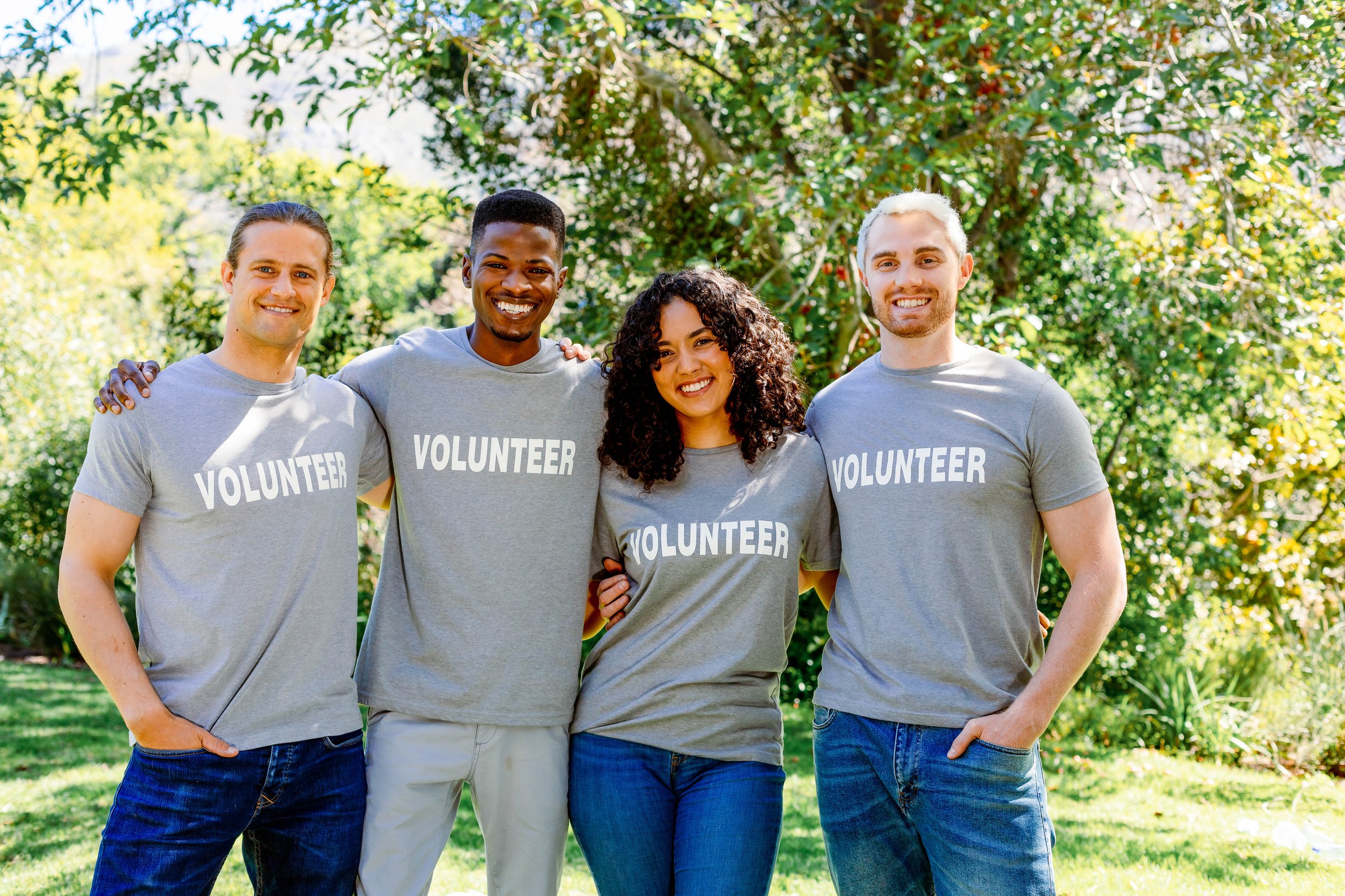 Four diverse volunteers wearing gray t-shirts that say 'VOLUNTEER' standing outdoors in a park, smiling with arms around each other, surrounded by green trees and sunlight.