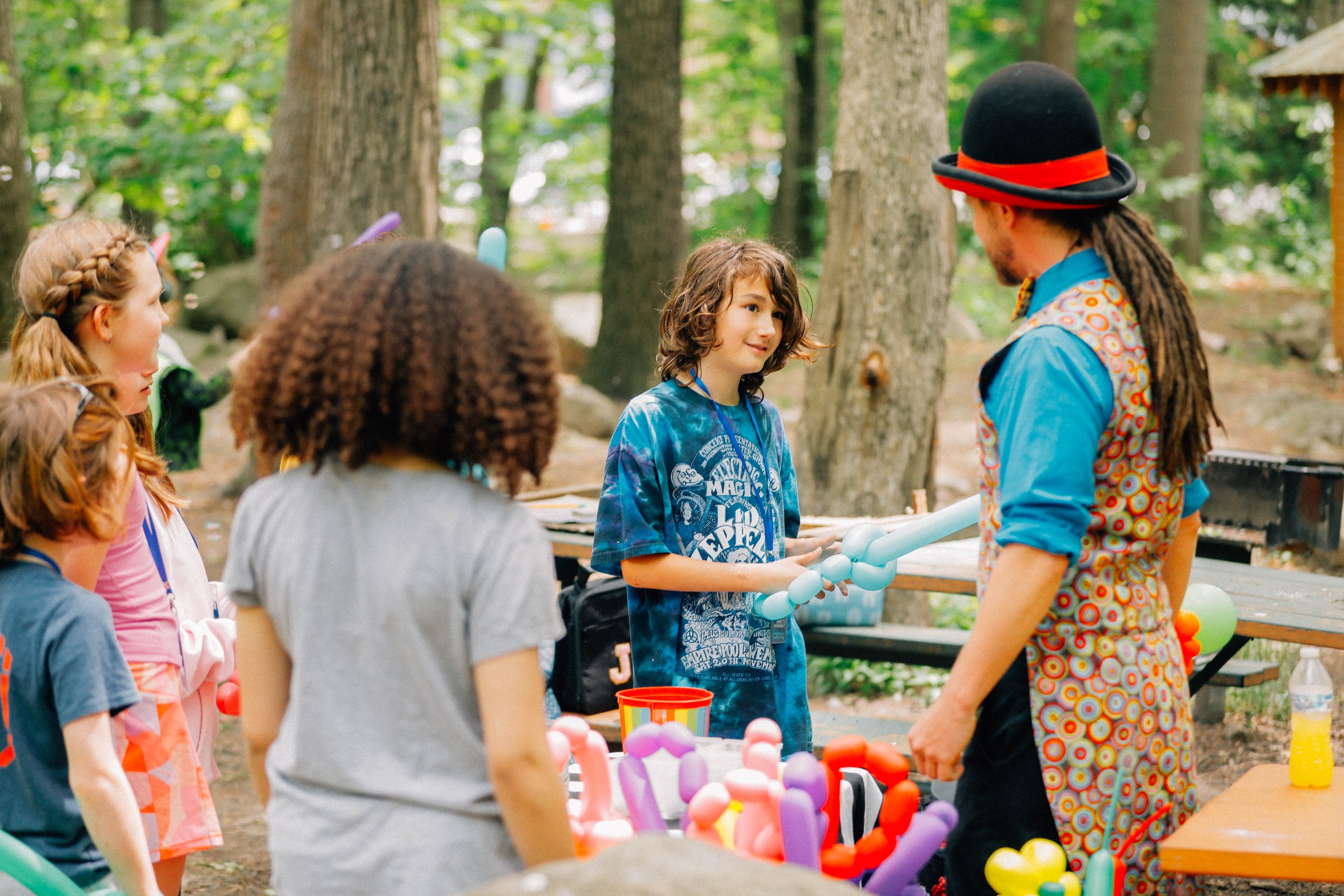 Children gathered around a clown at a birthday party in a park, with trees and natural surroundings in the background.