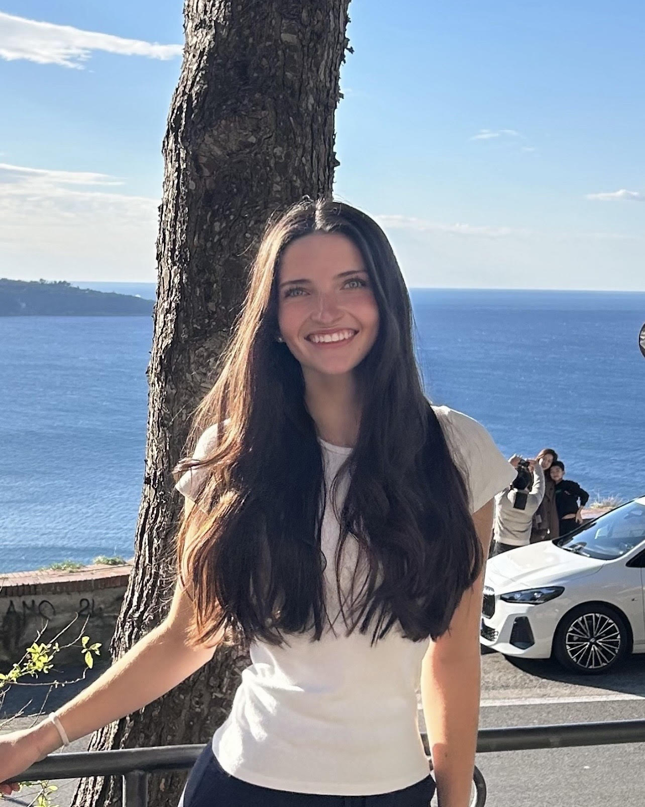 A young woman with long dark hair smiling in front of a scenic ocean view with a tree, parked cars, and people in the background.