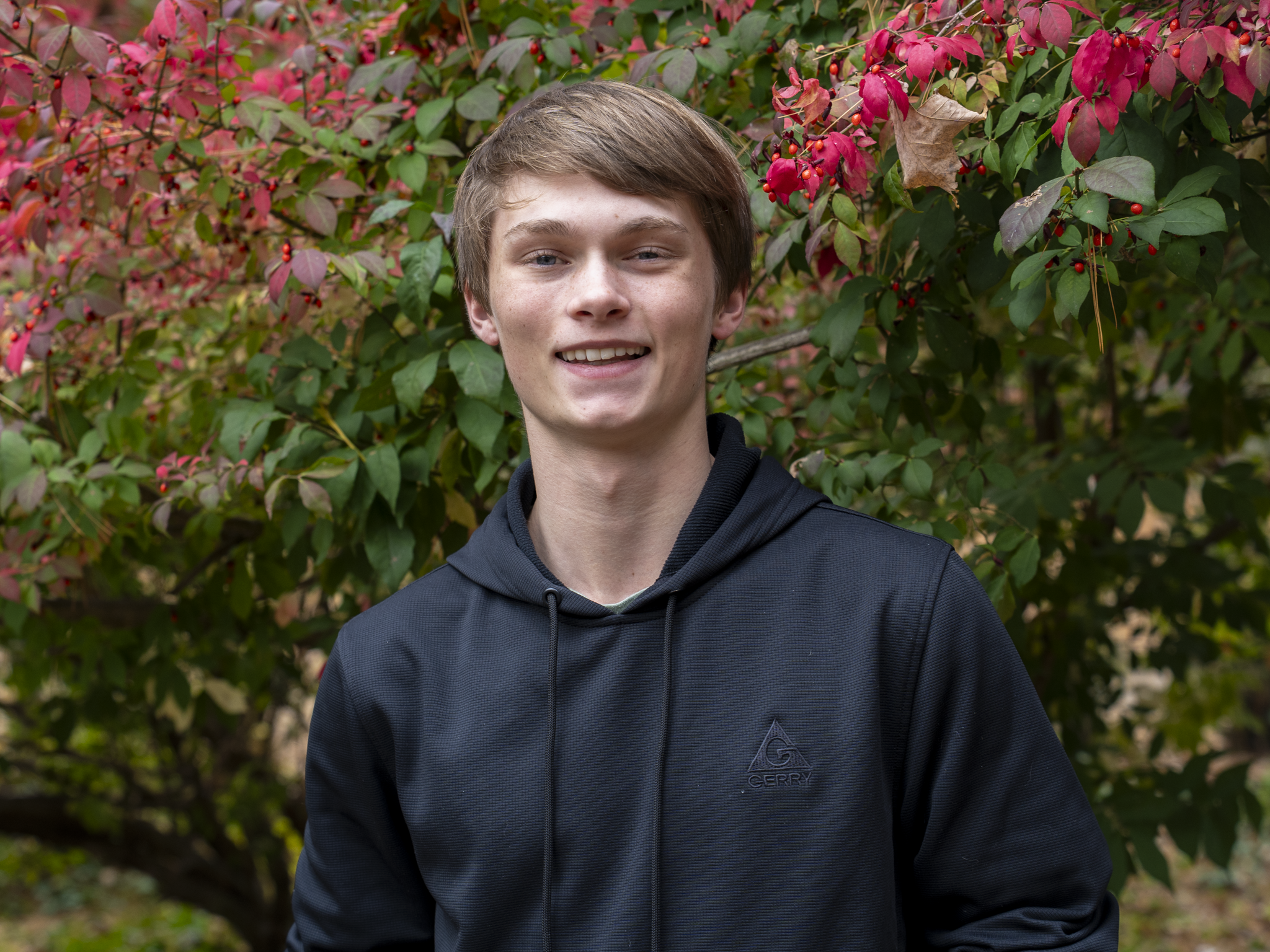 A young man with short brown hair, smiling and wearing a black jacket, standing outdoors in front of red and green foliage.