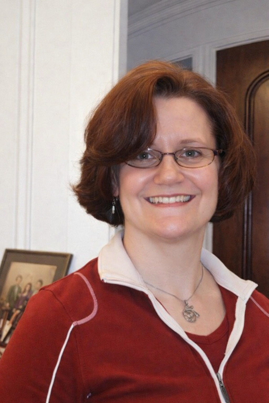 A woman with short, wavy, reddish-brown hair, wearing glasses, earrings, a necklace, and a red zip-up jacket with a white collar, smiling in an indoor setting.