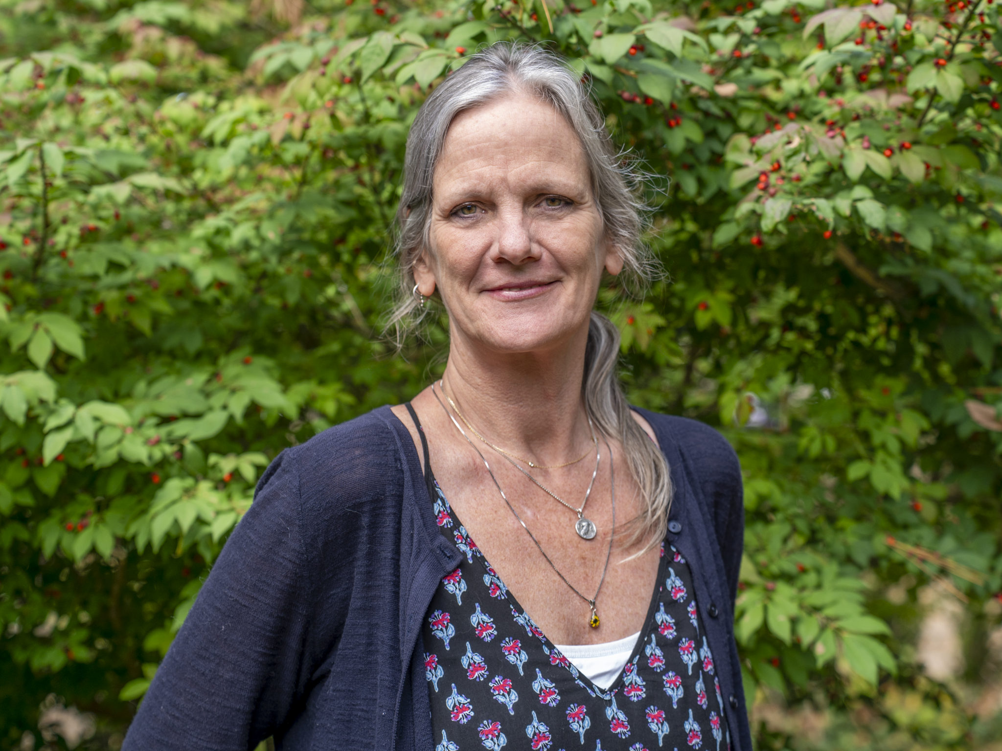 A middle-aged woman with gray hair, wearing a navy blue cardigan over a patterned dress, standing outdoors in front of green foliage with small red berries, smiling at the camera.