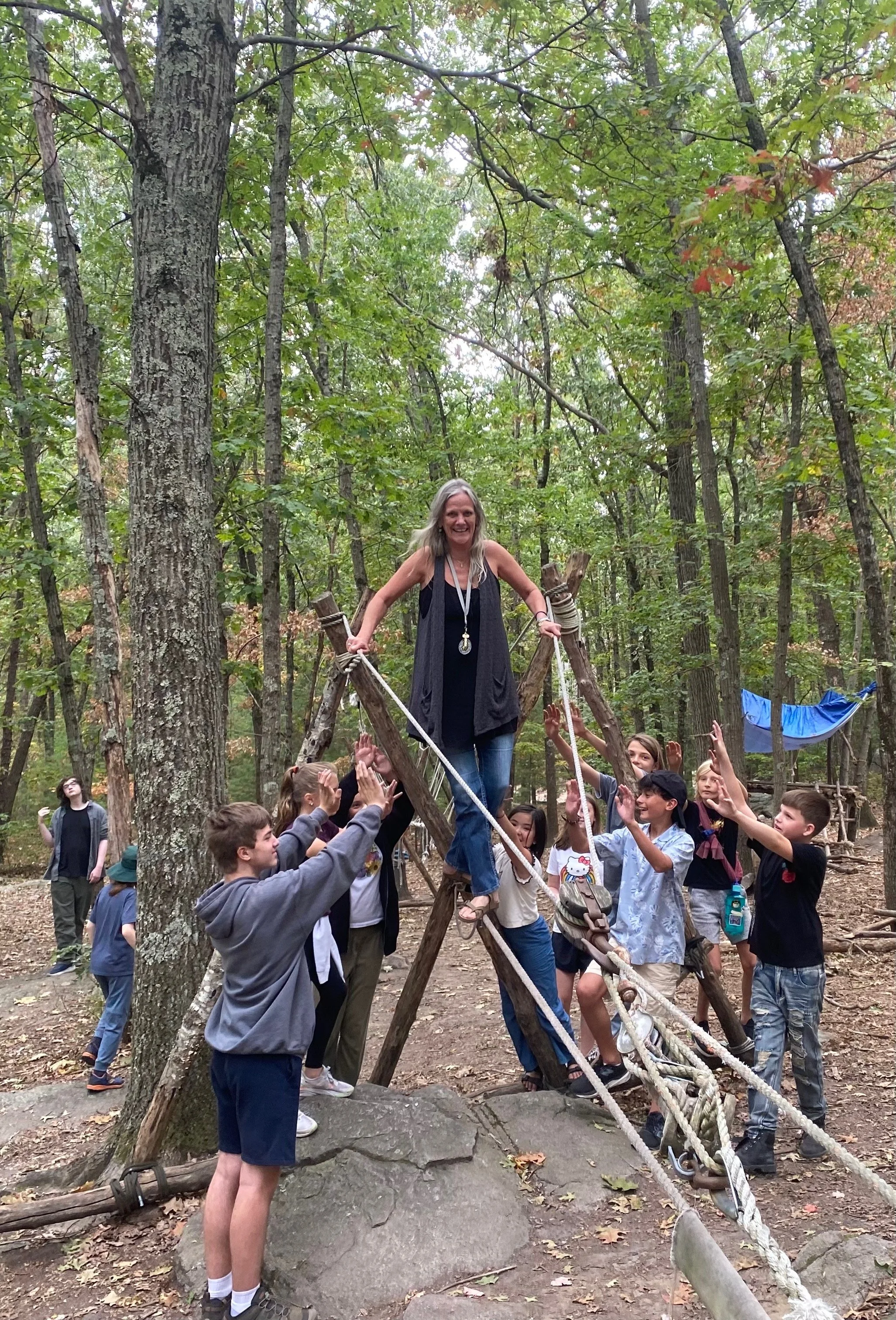 A woman standing on a wooden and rope structure in a forest, surrounded by children who are reaching up to her, with trees and a hammock in the background.