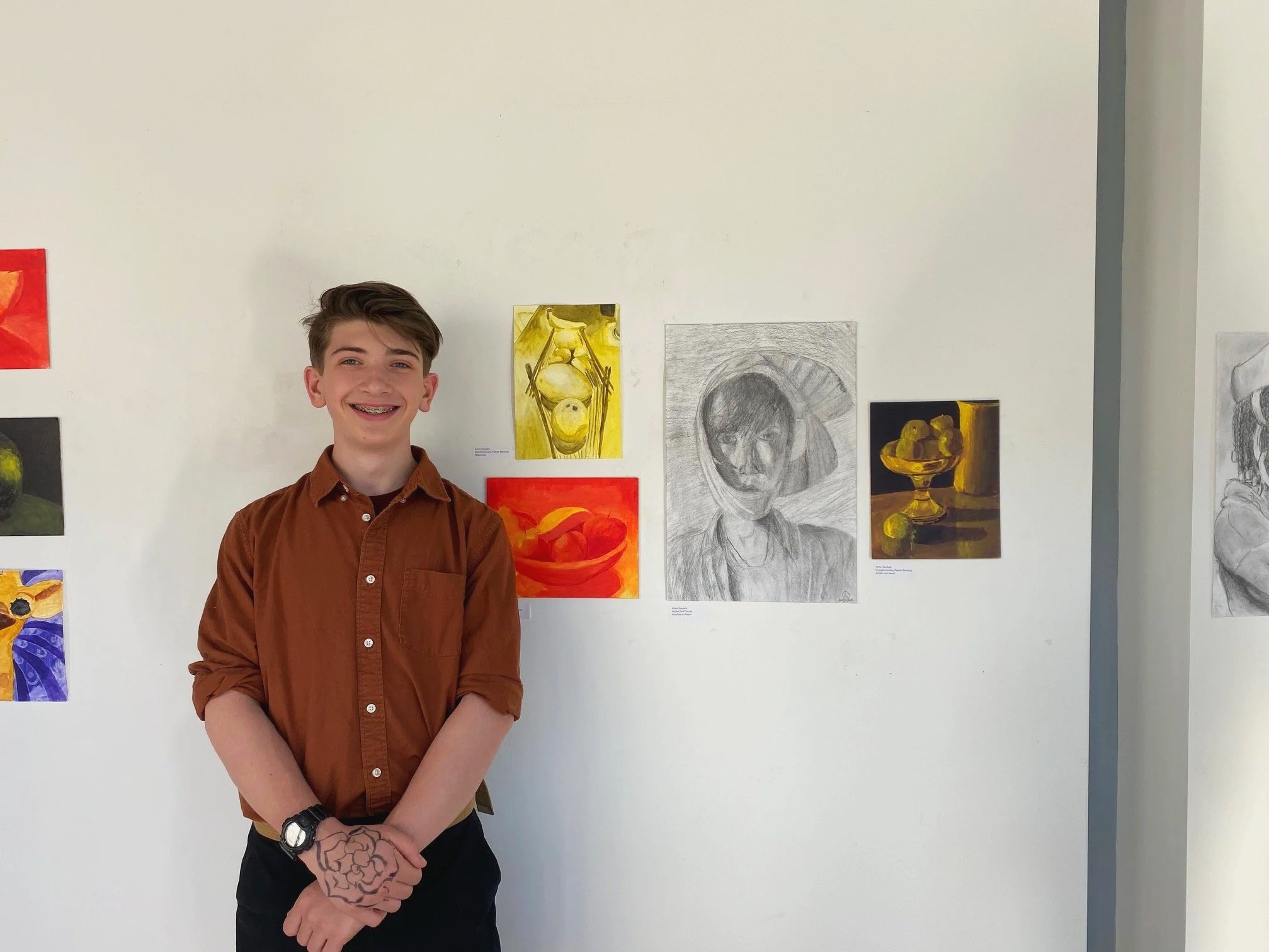 A young man in a brown shirt and black pants standing in front of an art gallery wall with various drawings and paintings, smiling at the camera.