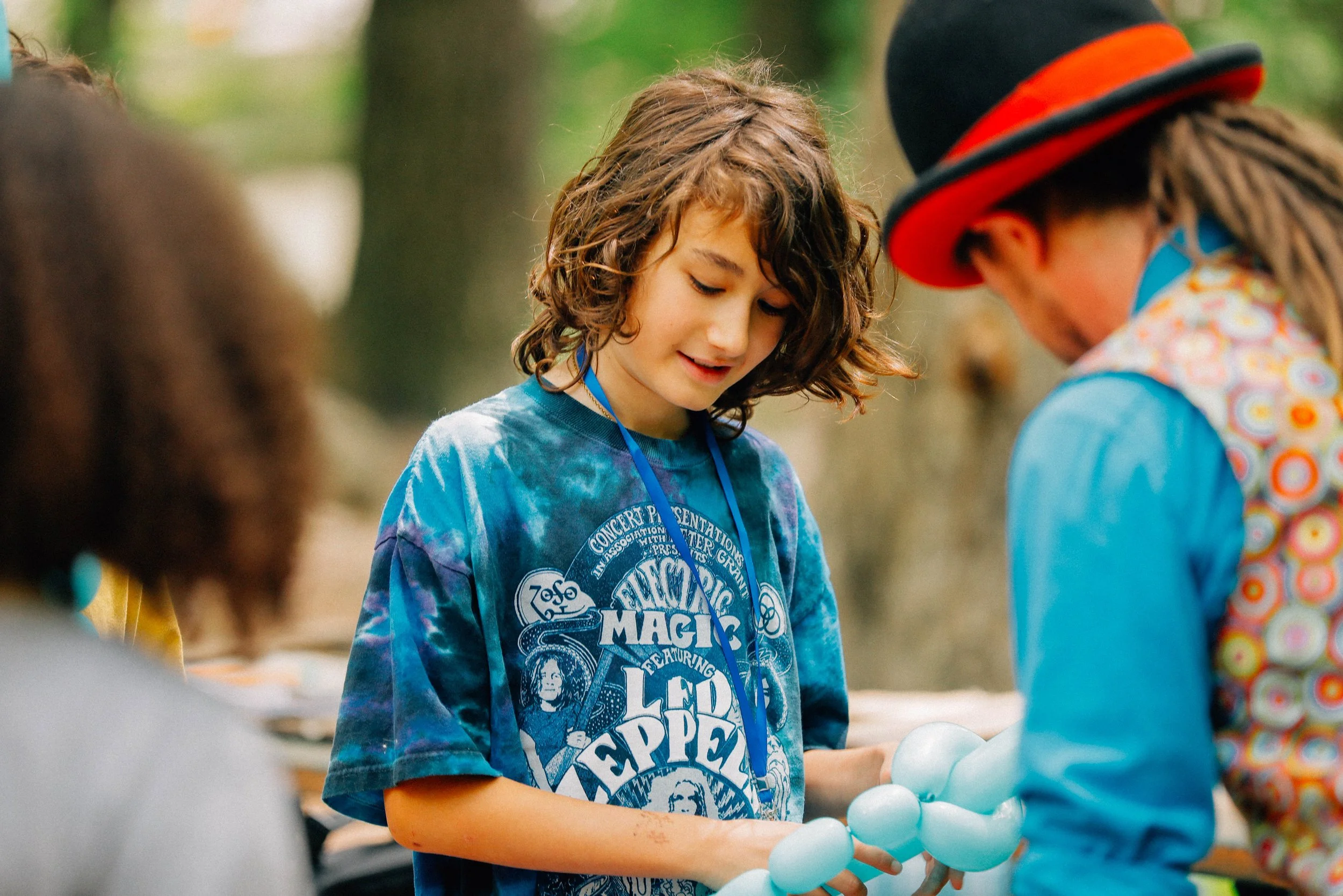 Young boy with curly hair wearing a Led Zeppelin T-shirt, holding a balloon sculpture, and a person in a colorful shirt and hat nearby, outdoors in a wooded area.