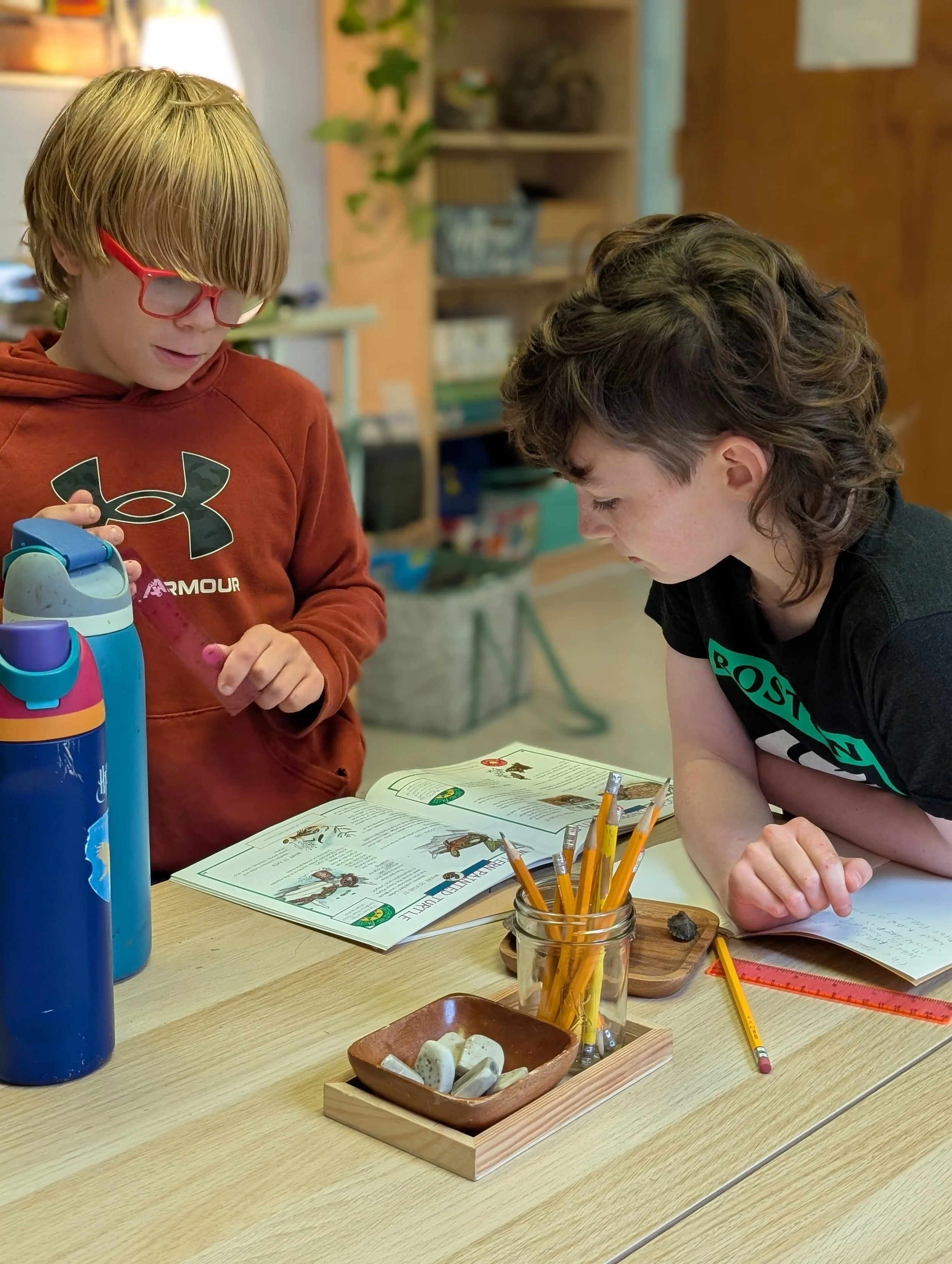 Two children at a table looking at a book, with school supplies and a water bottle on the table.