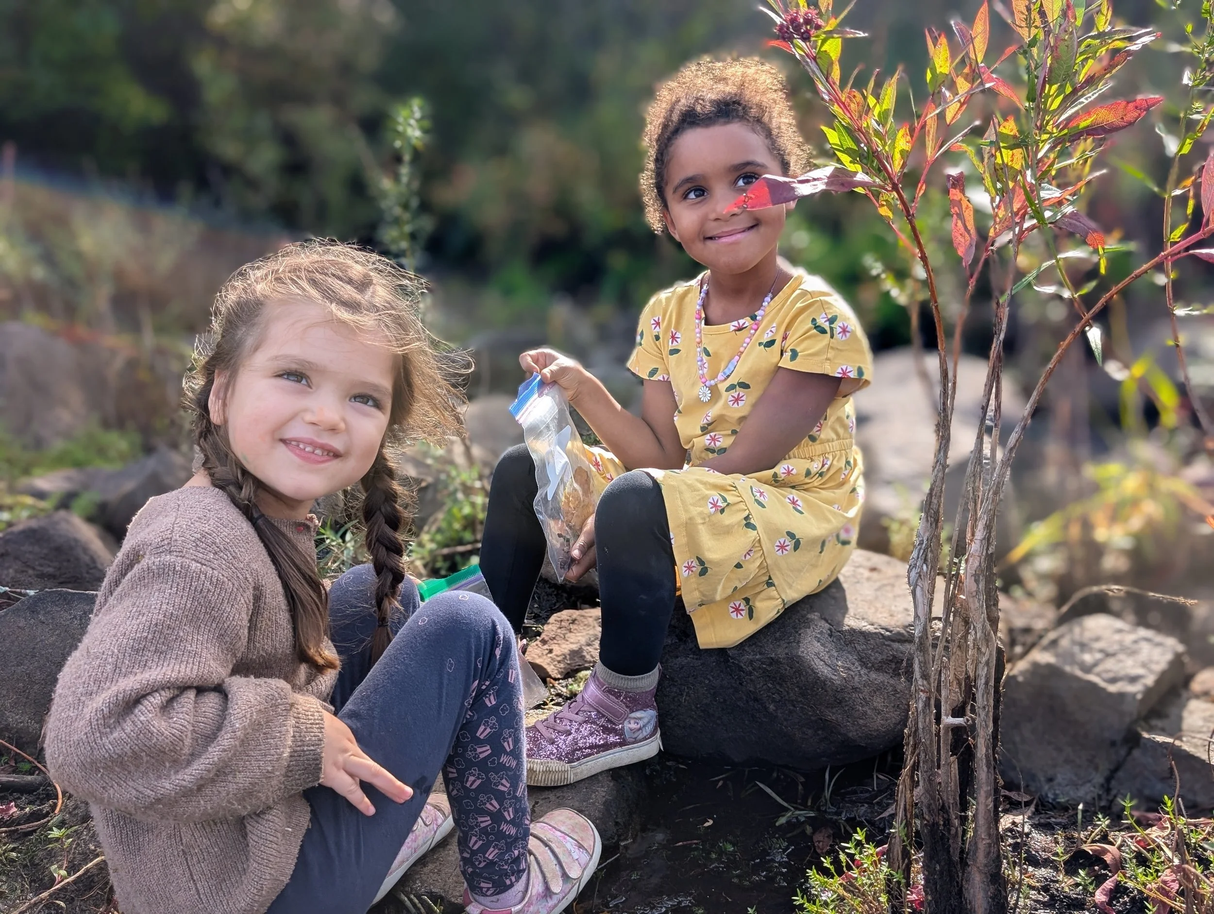 Two young girls sitting on rocks outdoors surrounded by plants and trees, one smiling at the camera and the other looking at the camera with a slight smile, holding a plastic bag.
