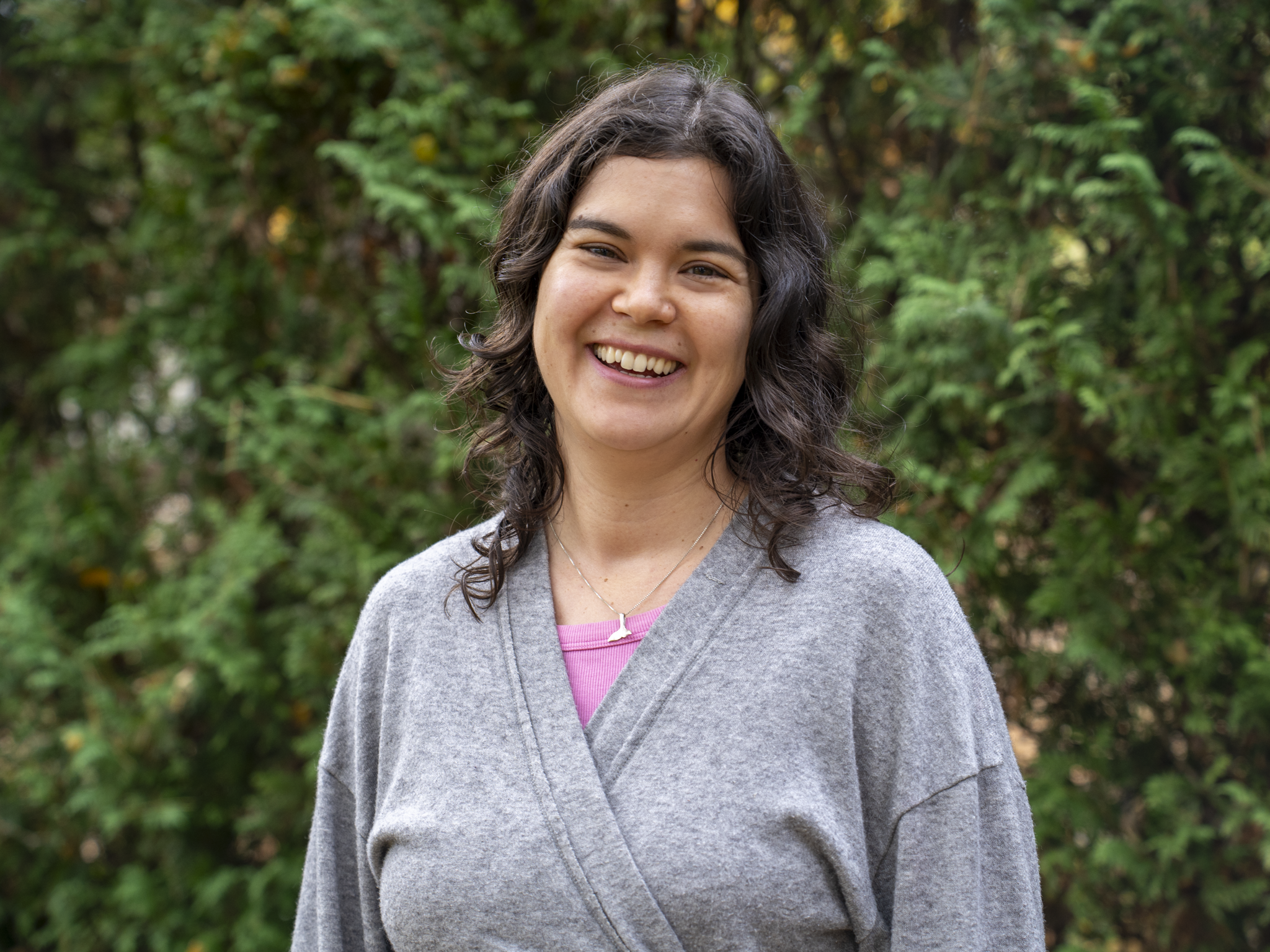 A woman with dark curly hair smiling outdoors in front of greenery.