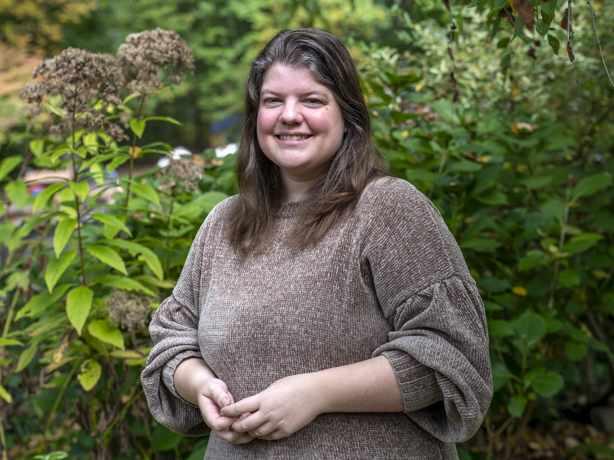 A woman with long brown hair smiling while standing among green plants and bushes.