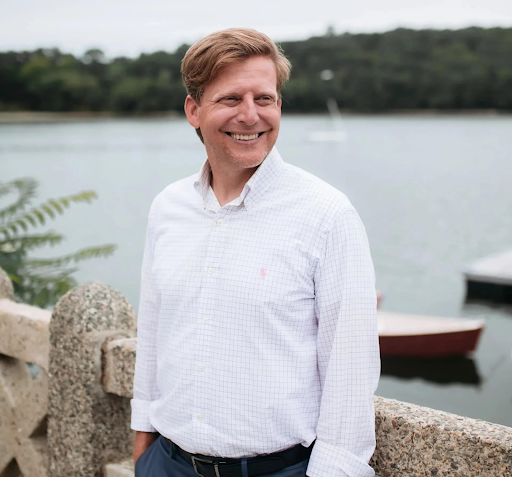 A smiling man in a white button-up shirt standing near a body of water with boats in the background and greenery on the far shore.