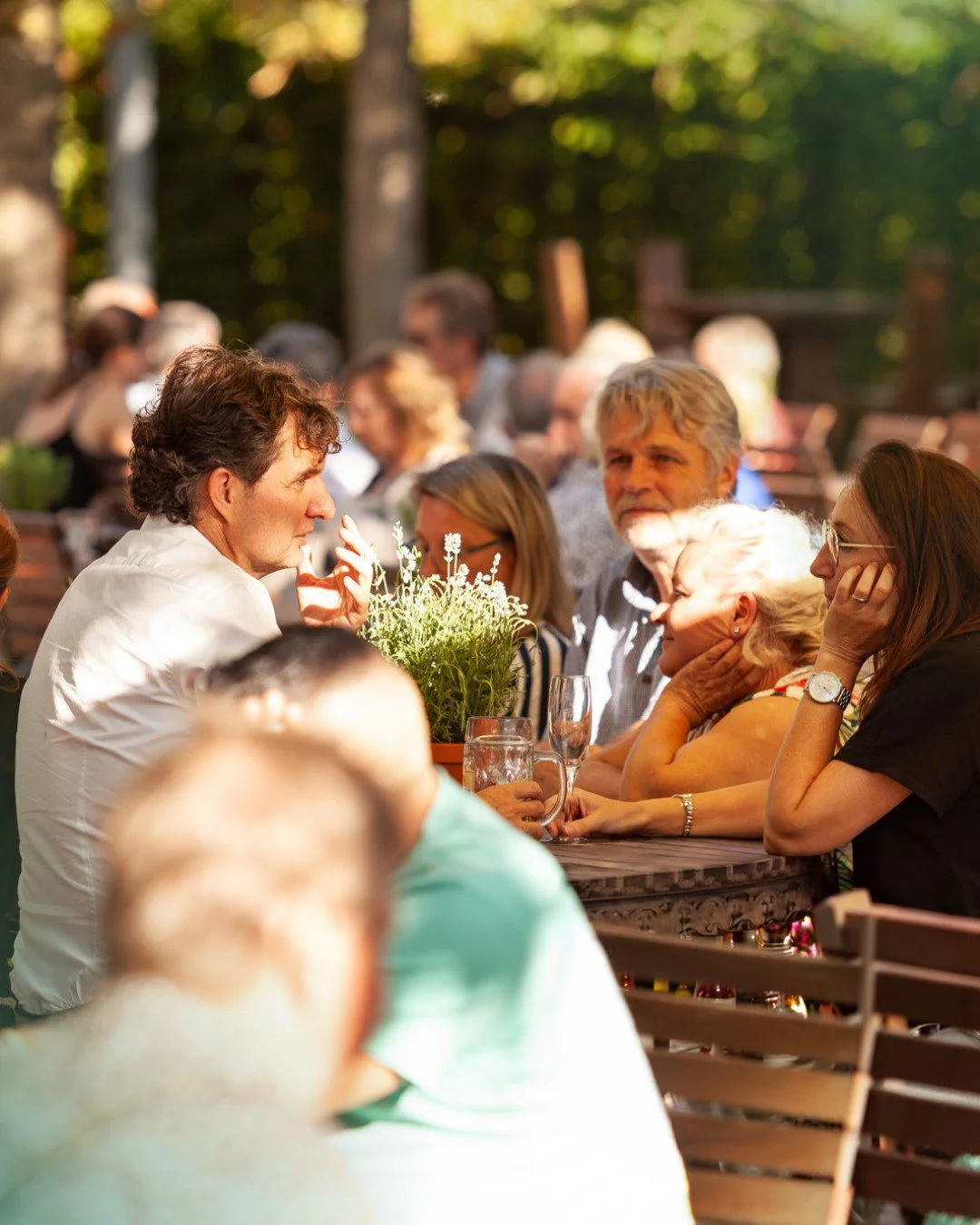  Eine sonnendurchflutete Biergartensituation mit Tischen an denen Menschen sitzen und sich unterhalten. 