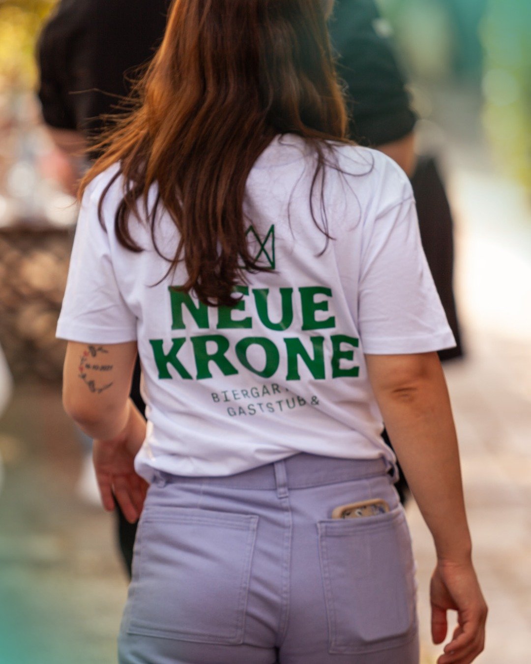  Eine Frau mit langen Haaren wendet der  Kamera den Rücken zu, auf ihrem T-Shirt sind Name und Logo der Neuen Krone zu erkennen. 