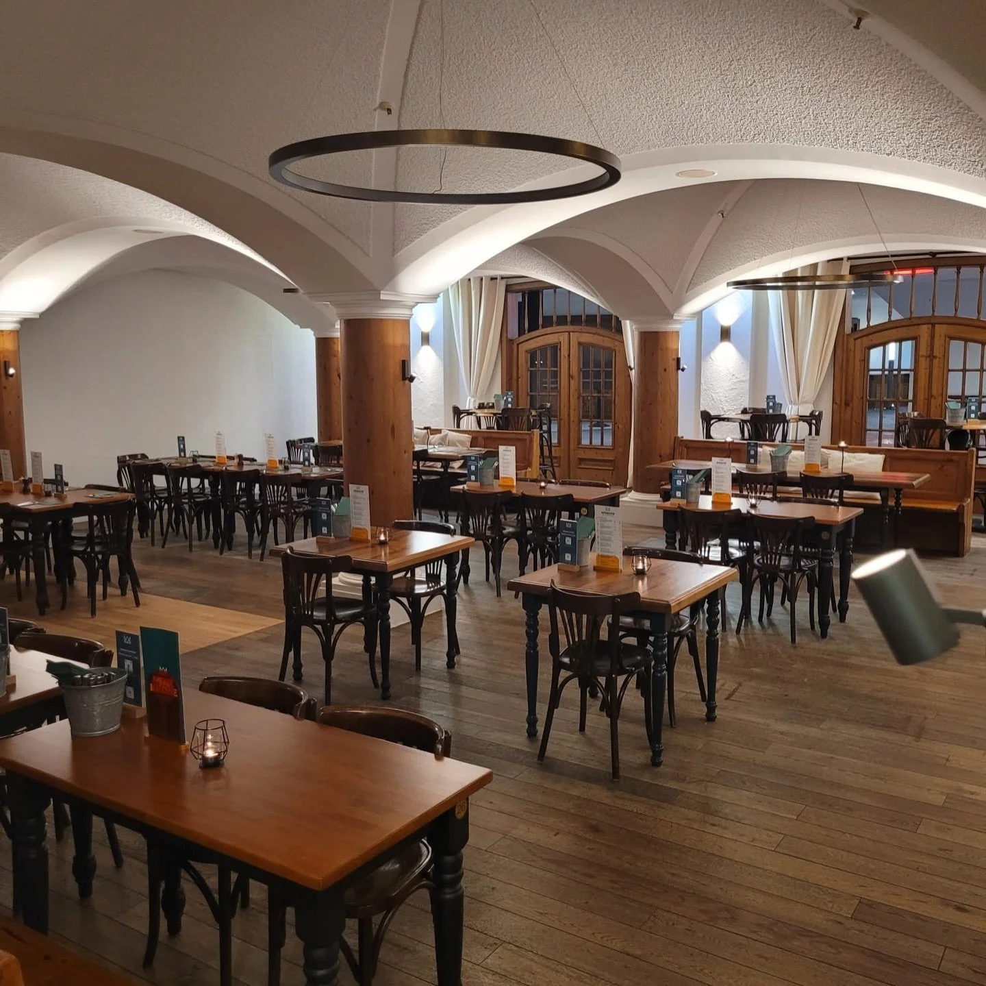 Innenansicht der Gaststube mit gemütlichen Holztischen und Stühlen, weißen Wölbedecken, gedecktem Licht, im Hintergrund Fenster mit Vorhängen.