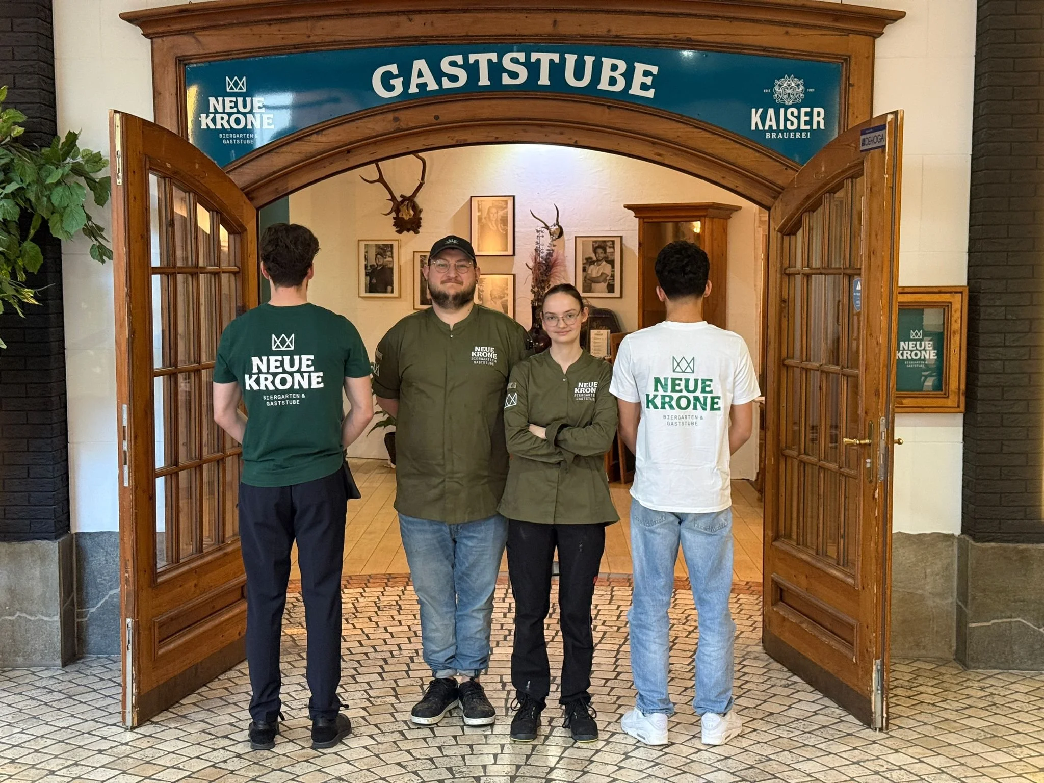  Im Eingang der Gastsube stehen 4 Personen aus dem Team, zwei Blicken in die Kamera, zwei wenden ihr den Rücken zu, auf ihren T-Shirts sind Name und Logo der Neuen Krone zu erkennen 