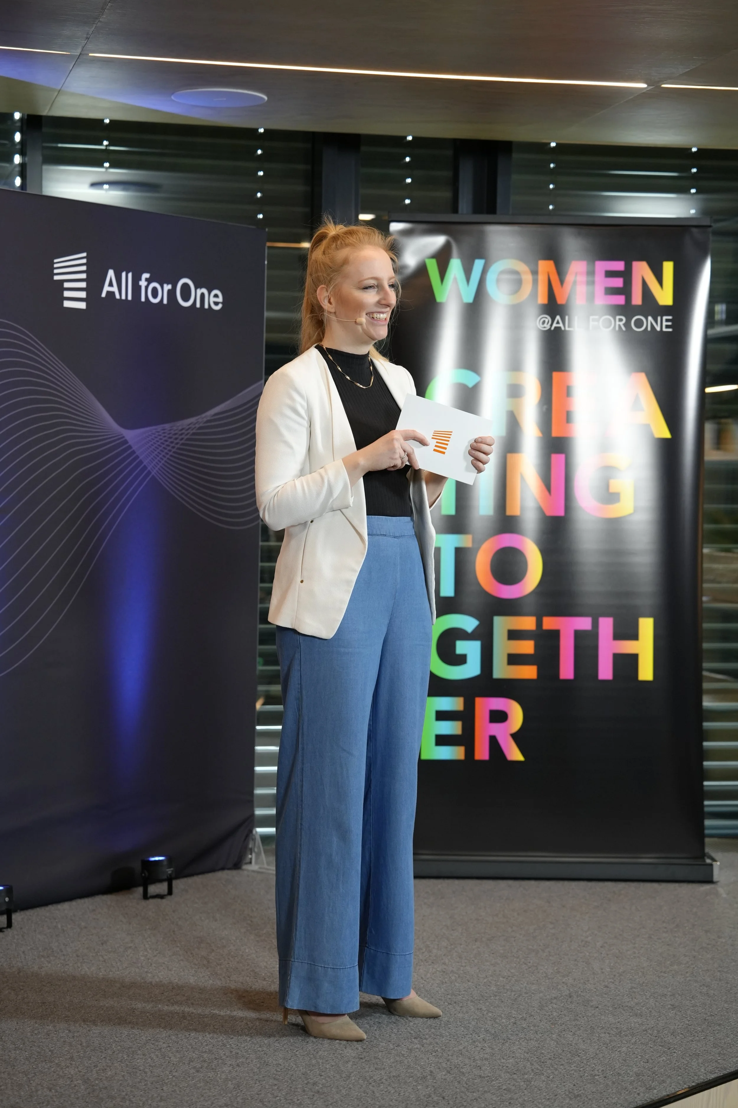 Eine junge Frau steht auf einer Bühne bei einer Veranstaltung, hält ein Whiteboard mit einem Logo und lächelt. Im Hintergrund sind Banner mit bunten Worten und Slogans wie „Women Creating Together“ und „All for One“.